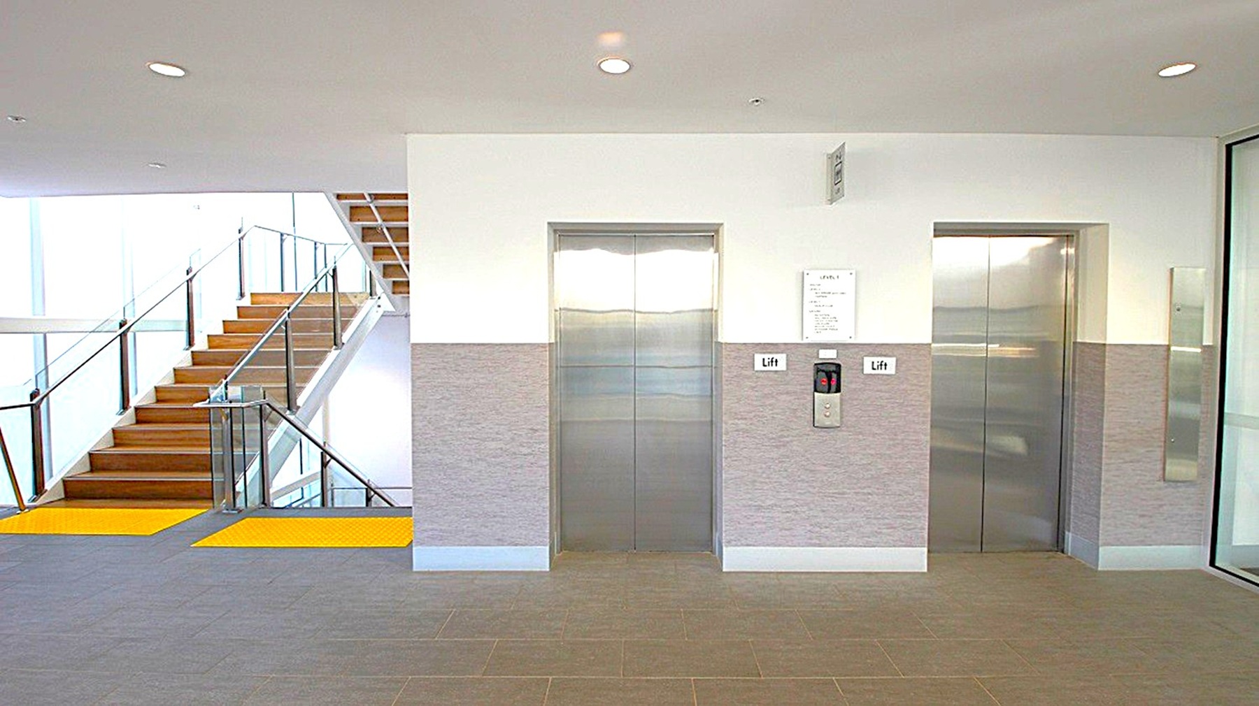 On the left, there is a staircase with wooden steps and glass railings, leading upwards. The middle features two lift doors with metallic finishes, set in a wall with a grey lower half and white upper half. A control panel and a sign labelled "Lift" are visible between the doors. On the right, a glass wall or door is partially visible. The floor is carpeted in a neutral tone, and the ceiling is white with recessed lighting.