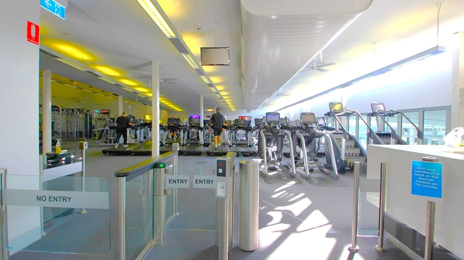 A gym interior with a row of treadmills on the right side, each equipped with digital screens. A person is using one of the treadmills. On the left, there are various gym machines and equipment. The entrance area in the foreground has gates marked "ENTRY" and "NO ENTRY." The ceiling is lined with fluorescent lights, and large windows on the right allow natural light to illuminate the space. The floor is carpeted in a neutral colour.