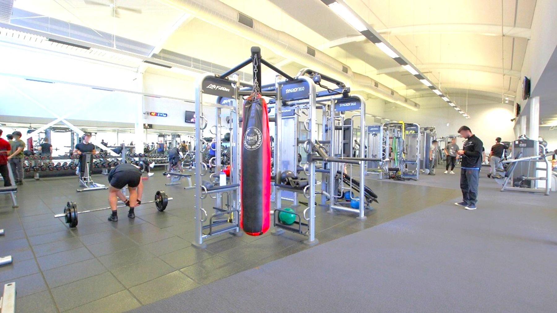 A spacious gym with various exercise equipment. In the centre, there's a large multi-functional machine with a red and black punching bag attached. To the left, a person is bending over a barbell, while others are using weights in the background. On the right, a person is standing, possibly checking a device. The floor is covered with dark rubber mats, and the ceiling is high with fluorescent lighting. The gym is well-lit with natural light coming through large windows.