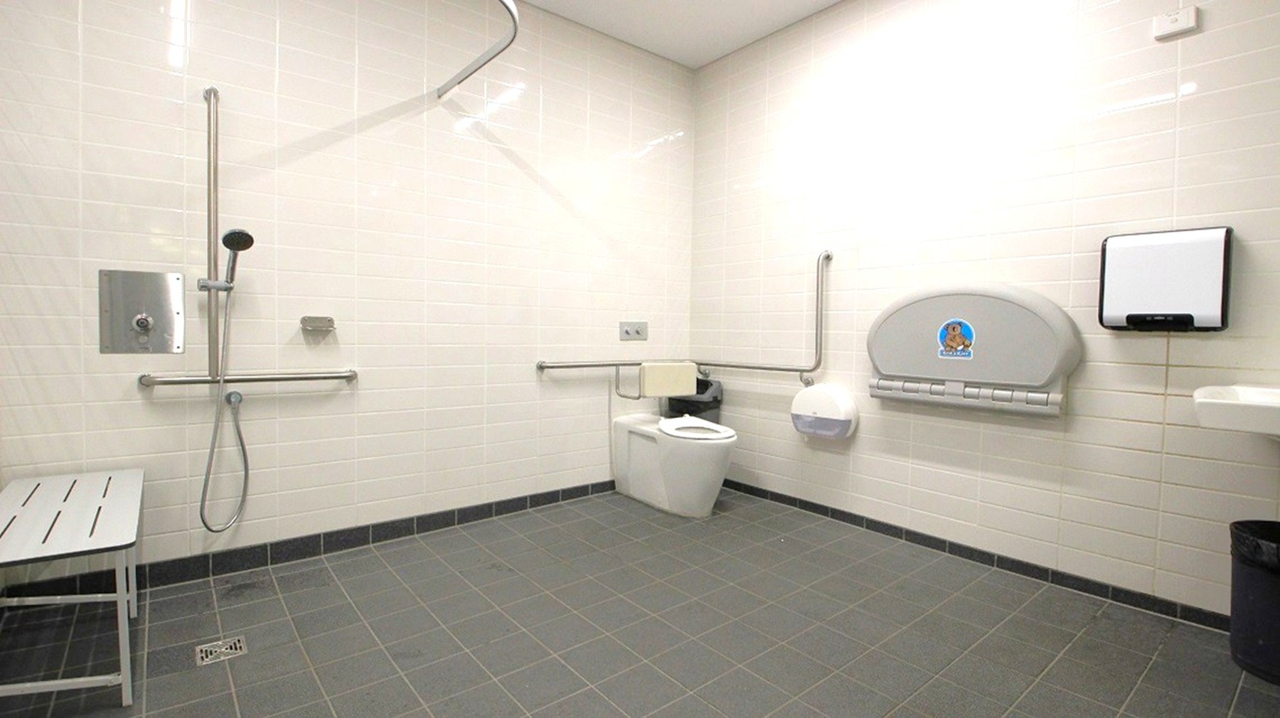 A spacious, accessible bathroom with light-coloured tiled walls and a grey tiled floor. On the left, there is a shower area with a handheld showerhead and a metal grab bar. A bench is positioned nearby. On the right, a toilet with support rails is visible, along with a baby changing station mounted on the wall. A paper towel dispenser and a waste bin are also present. The ceiling is plain and light-coloured.