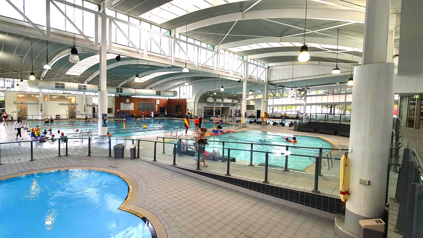 An indoor swimming pool complex with multiple pools. On the left, a smaller, round pool is visible with a tiled edge. In the centre, a larger rectangular pool is filled with people swimming and engaging in activities. The right side shows another pool area with people and a lifeguard. The ceiling is high with large windows and hanging lights, supported by white beams and columns. The floor is tiled, and there are railings separating the pool areas.