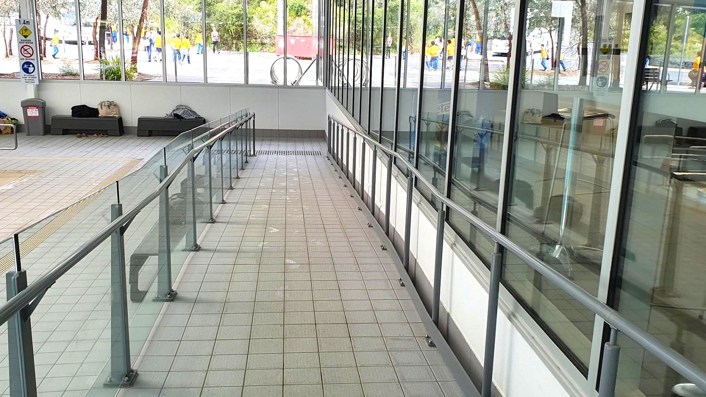 A tiled ramp with metal railings on both sides leads downwards. On the left, there are benches with bags placed on them and a rubbish bin nearby. Large windows on the right provide a view of people walking outside, wearing yellow shirts. The floor is tiled in a light grey and green pattern. Signs are visible on the wall to the left, and there are plants outside the windows.