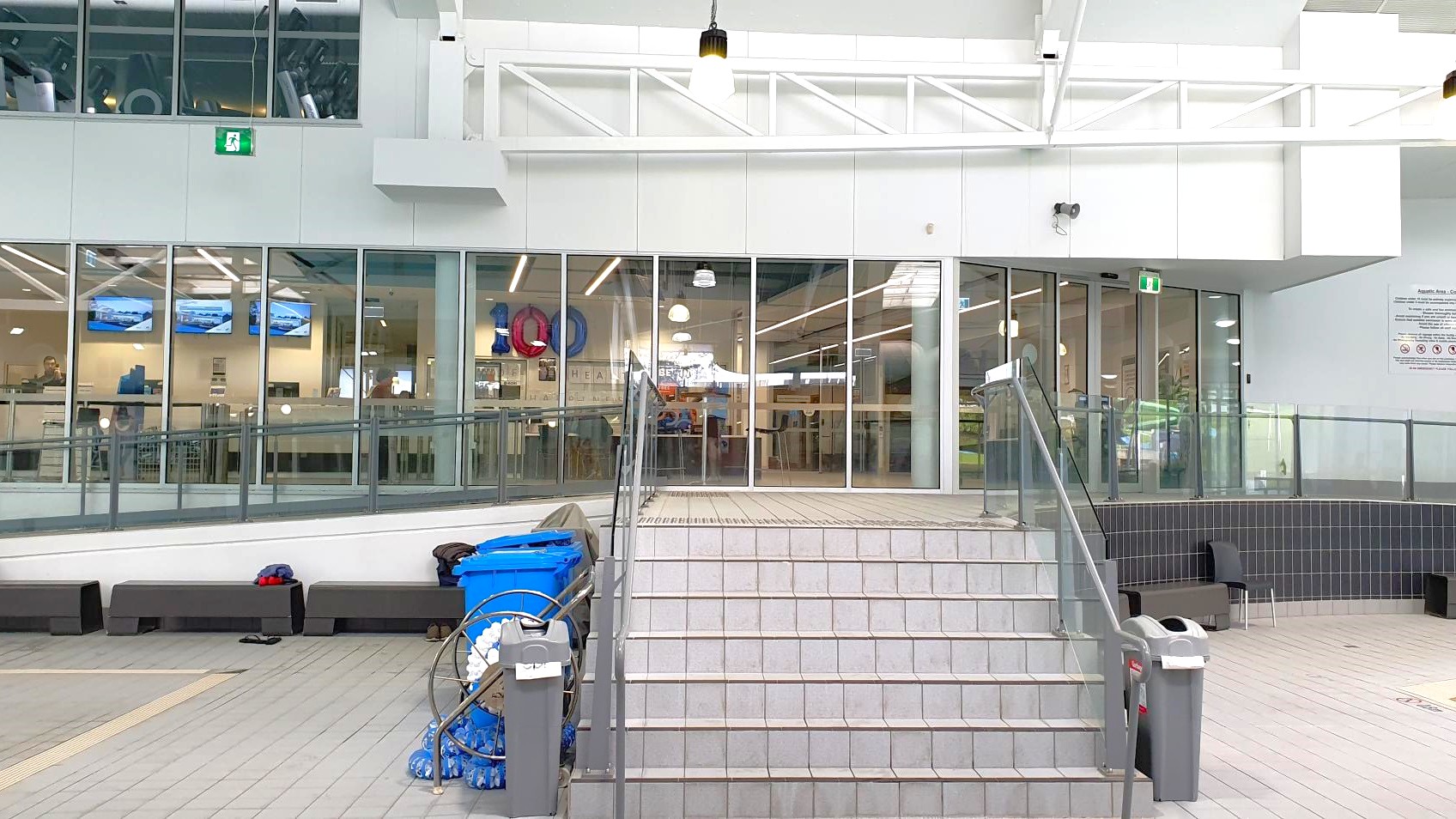 A set of grey tiled stairs leads up to a glass-walled interior space. The glass panels reveal a room with several screens displaying images and a few people inside. On the left, there are blue bins and cleaning equipment. The ceiling is white with exposed beams and hanging lights. The floor is tiled in grey, and there are black benches along the wall. A green exit sign is visible above the glass doors.