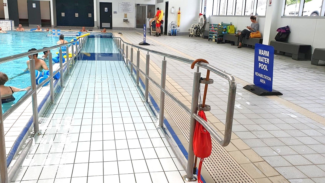 A swimming pool with a ramp on the left, featuring several people in the water using flotation devices. The right side has a tiled floor with a handrail and a red flotation device attached. A sign reads "Warm Water Pool Rehabilitation Area." In the background, a lifeguard stands near a wall, and a person sits on a bench reading. Large windows line the right wall, allowing natural light to enter.