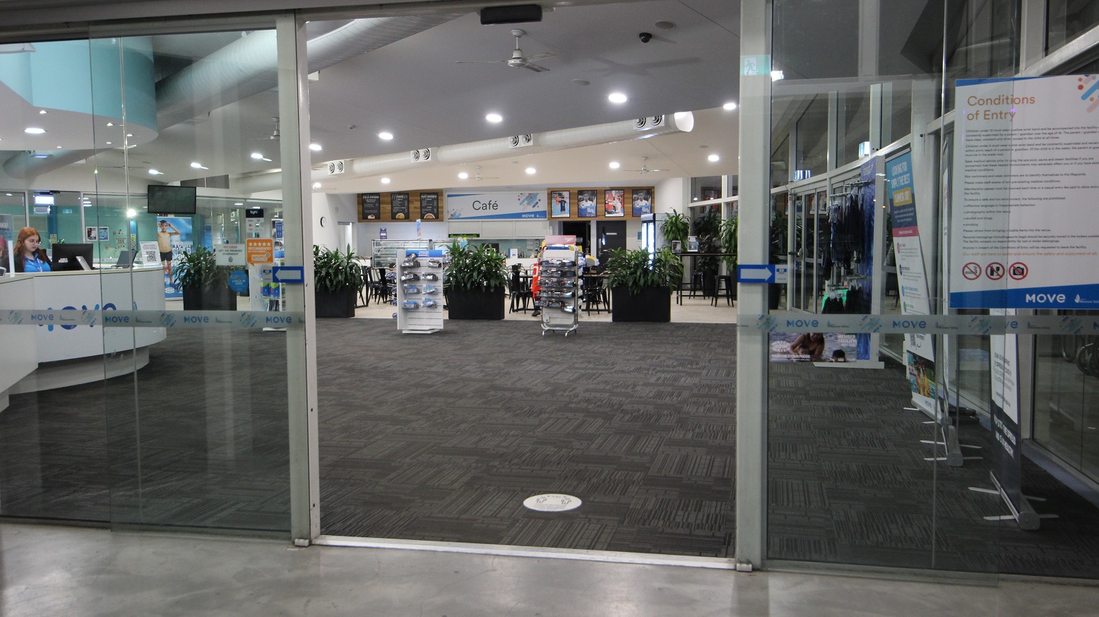 The entrance to the building is via an automatic glass sliding door, which displays a poster detailing entry conditions. Inside, the interior includes a grey carpet and white ceiling equipped with LED lighting and large air conditioning vents. To the left is a reception desk with a white facade, adjacent to a retail display featuring goggles. A large retractable banner stands to the right. In the centre, a large planter box filled with indoor plants is aligned, with a cafe situated behind it.