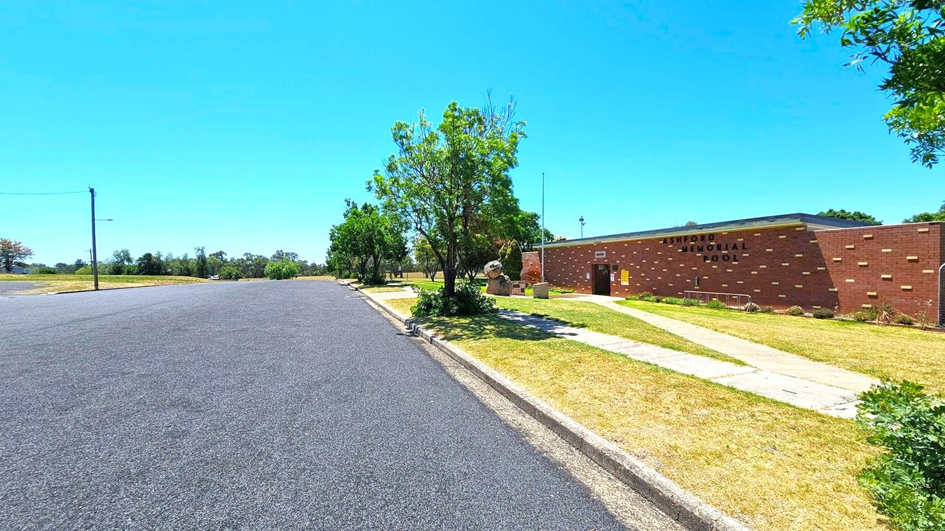 A quiet street with a wide, empty road on the left. On the right, a brick building with the words "ASHFORD MEMORIAL POOL" is visible. A pathway leads to the entrance, surrounded by a grassy area with a few trees and shrubs. A single streetlamp stands on the left side of the road. The background features more trees and open space.