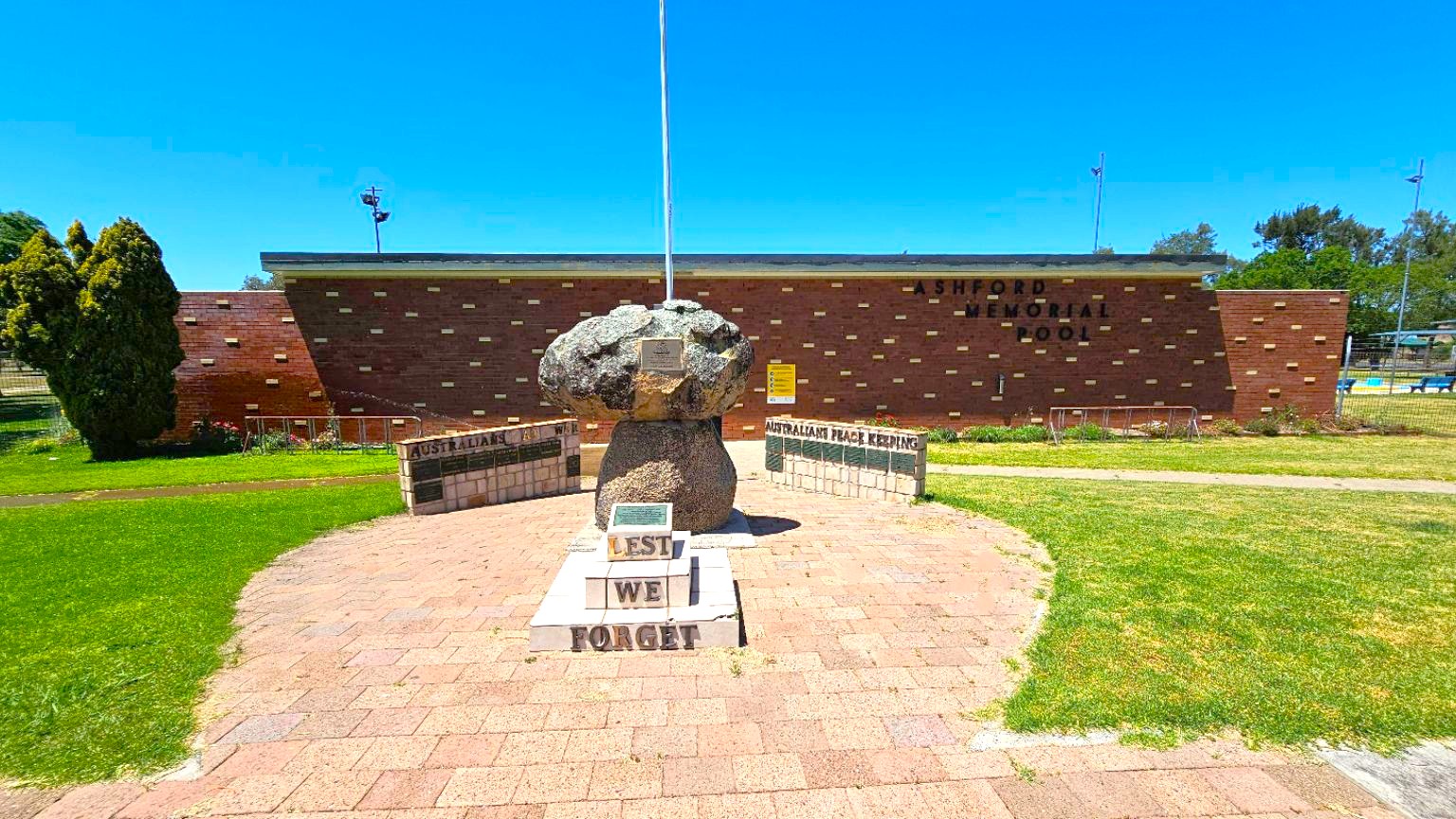 A memorial with a stone monument and a flagpole in the centre, surrounded by plaques. The base of the monument has the words "LEST WE FORGET" inscribed. The background features a brick building with the words "ASHFORD MEMORIAL POOL" on it. To the left, there is a tree and some grass, while to the right, there is a fenced area with more grass. The ground is paved with red and grey bricks.