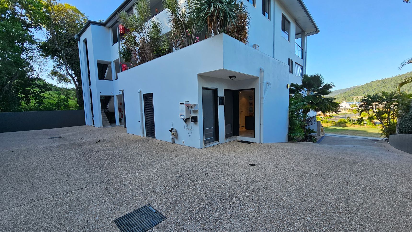 A modern white building with multiple floors is on the left, featuring a staircase and several windows. The ground is paved with a textured surface. On the right, there is a driveway leading downwards, bordered by lush greenery and palm trees. In the background, hills and a clear sky are visible. The building has a small garden with plants on the first-floor balcony.