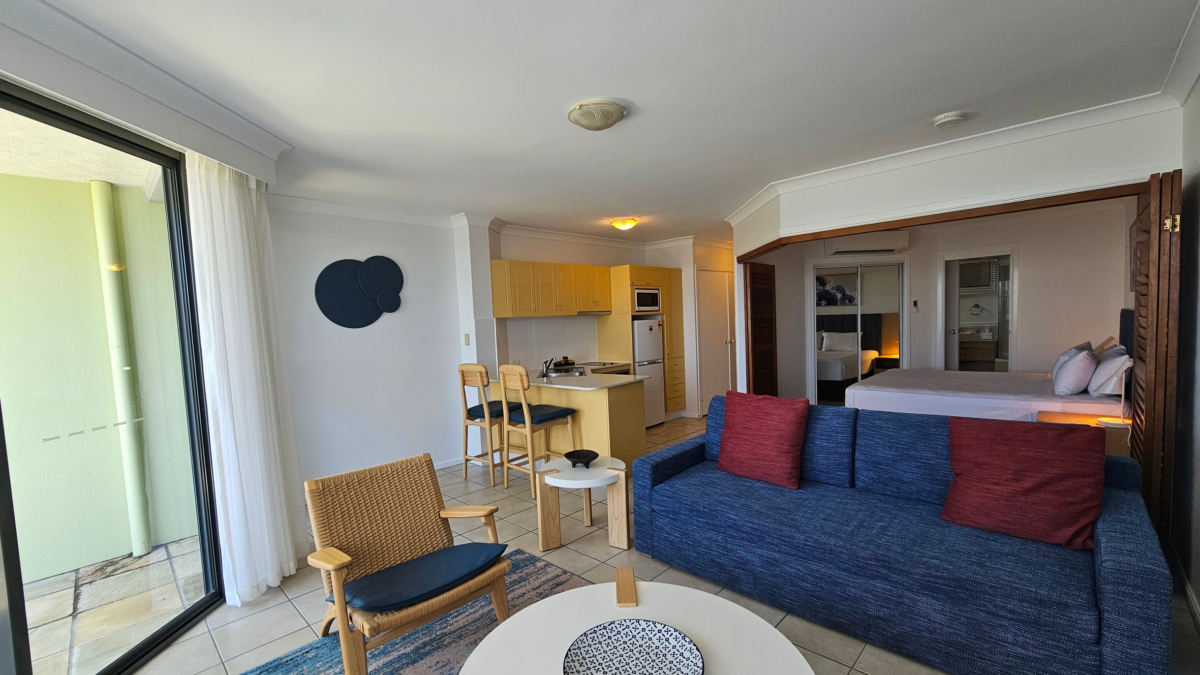 A modern living space with a sliding glass door on the left, leading to a balcony. Inside, there's a wicker chair and a blue sofa with red cushions. A round coffee table with a decorative bowl sits in front. The kitchen area is in the background, featuring light wood cabinets, a small island with two bar stools, and appliances. On the right, a bedroom area with a bed and mirrored wardrobe is partially visible. The floor is tiled, and the ceiling has recessed lighting.