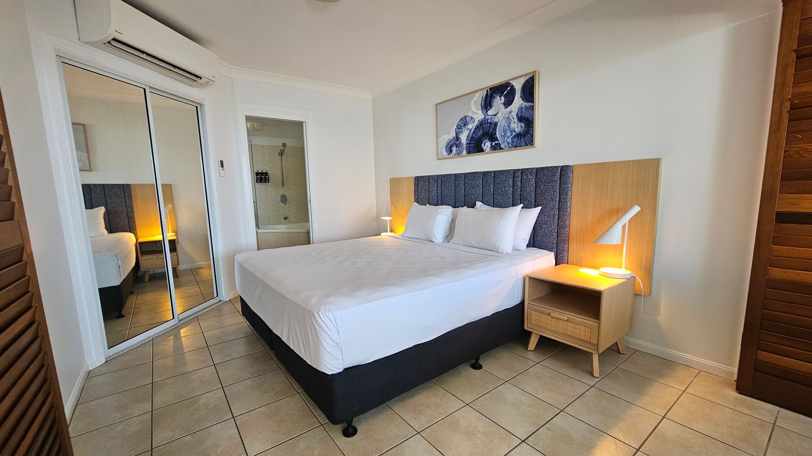 A bedroom with a large bed in the centre, featuring a blue and wood headboard. On the right, a wooden bedside table with a modern lamp. Above the bed, a framed abstract artwork. To the left, mirrored sliding doors reflect the room, with an air conditioning unit above. The floor is tiled, and a glimpse of a bathroom is visible through an open door.
