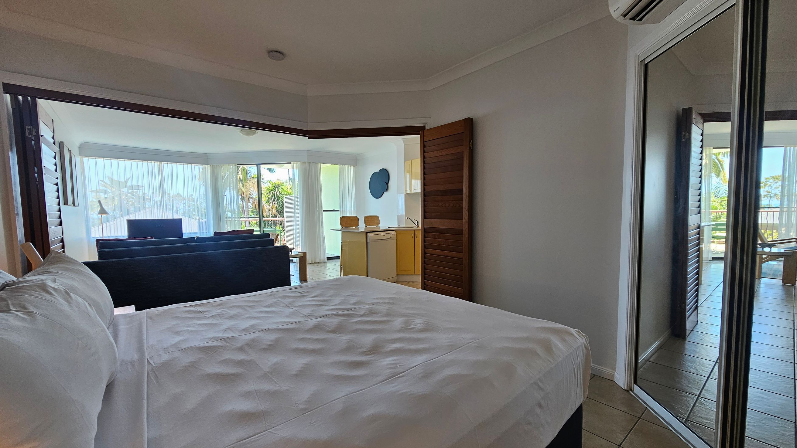 A bedroom with a neatly made bed on the left, featuring white linens. To the right, a mirrored wardrobe reflects part of the room and a glimpse of the outside. In the middle, an open doorway leads to a living area with a dark sofa, a small table, and a kitchen with wooden cabinets and two bar stools. Large windows with sheer curtains reveal palm trees and a balcony outside. The floor is tiled, and an air conditioning unit is mounted on the wall.