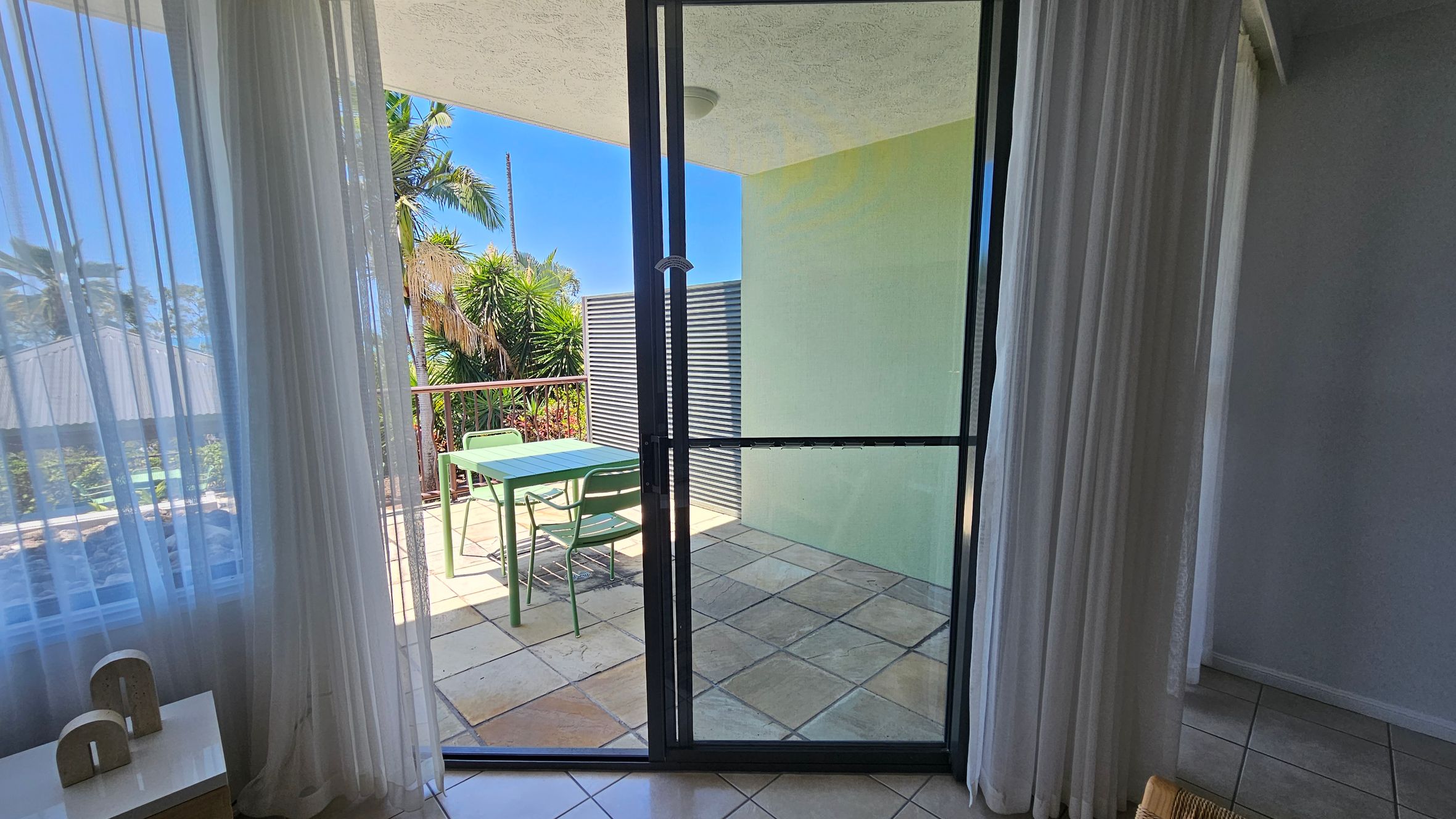 A room with sheer white curtains on the left, partially open to reveal a sliding glass door leading to a balcony. The balcony has a green table and chairs on a tiled floor. In the background, there are lush green plants and palm trees. The interior floor is tiled, and a small white table with decorative items is visible inside. The ceiling is plain and light-coloured.