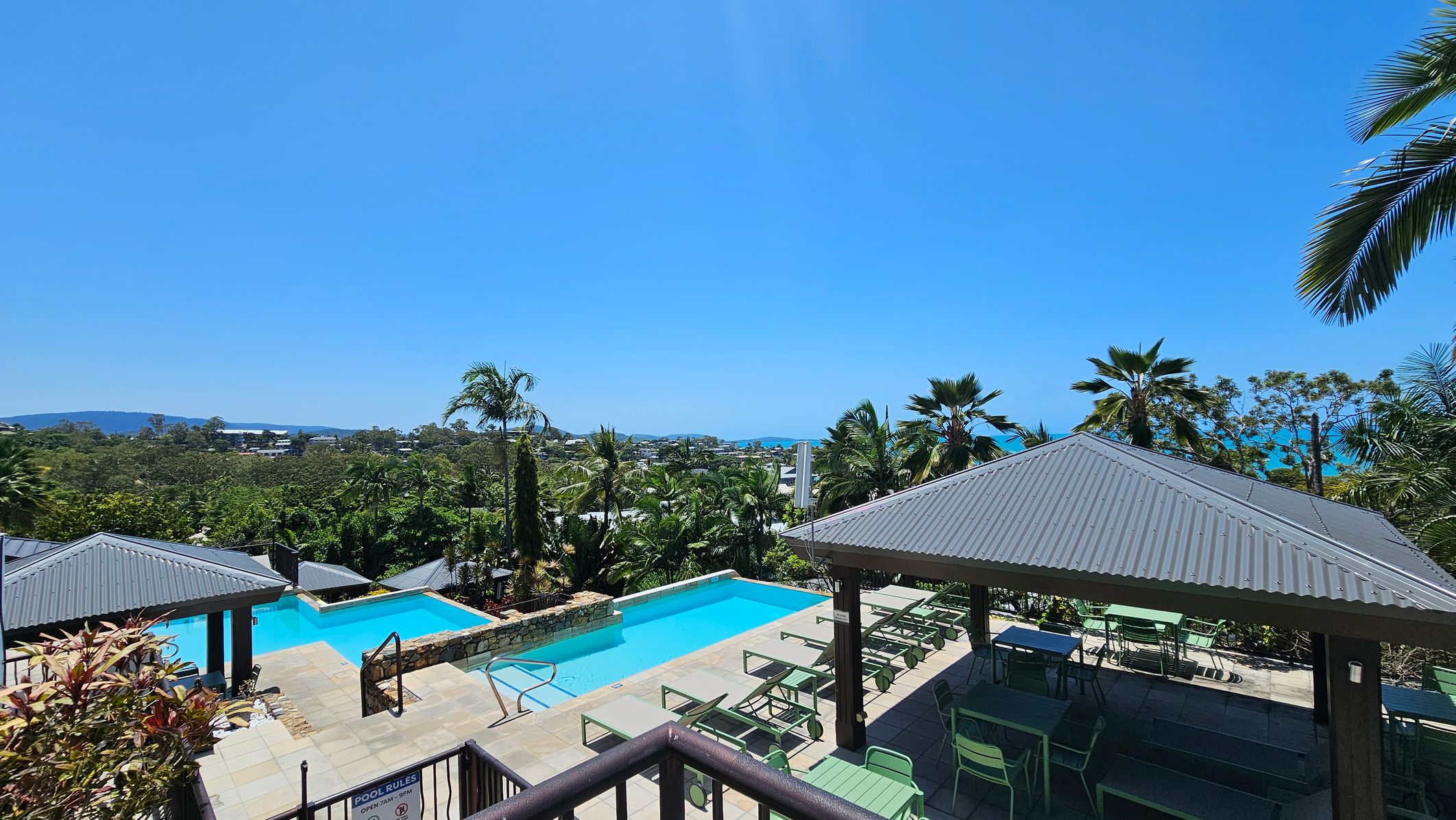 A swimming pool area with a clear blue pool in the centre, surrounded by lush green palm trees. On the right, there is a shaded seating area with green tables and chairs under a corrugated metal roof. On the left, another similar structure is partially visible. In the background, there are more trees and a view of distant hills. The foreground features a railing and a sign with pool rules.