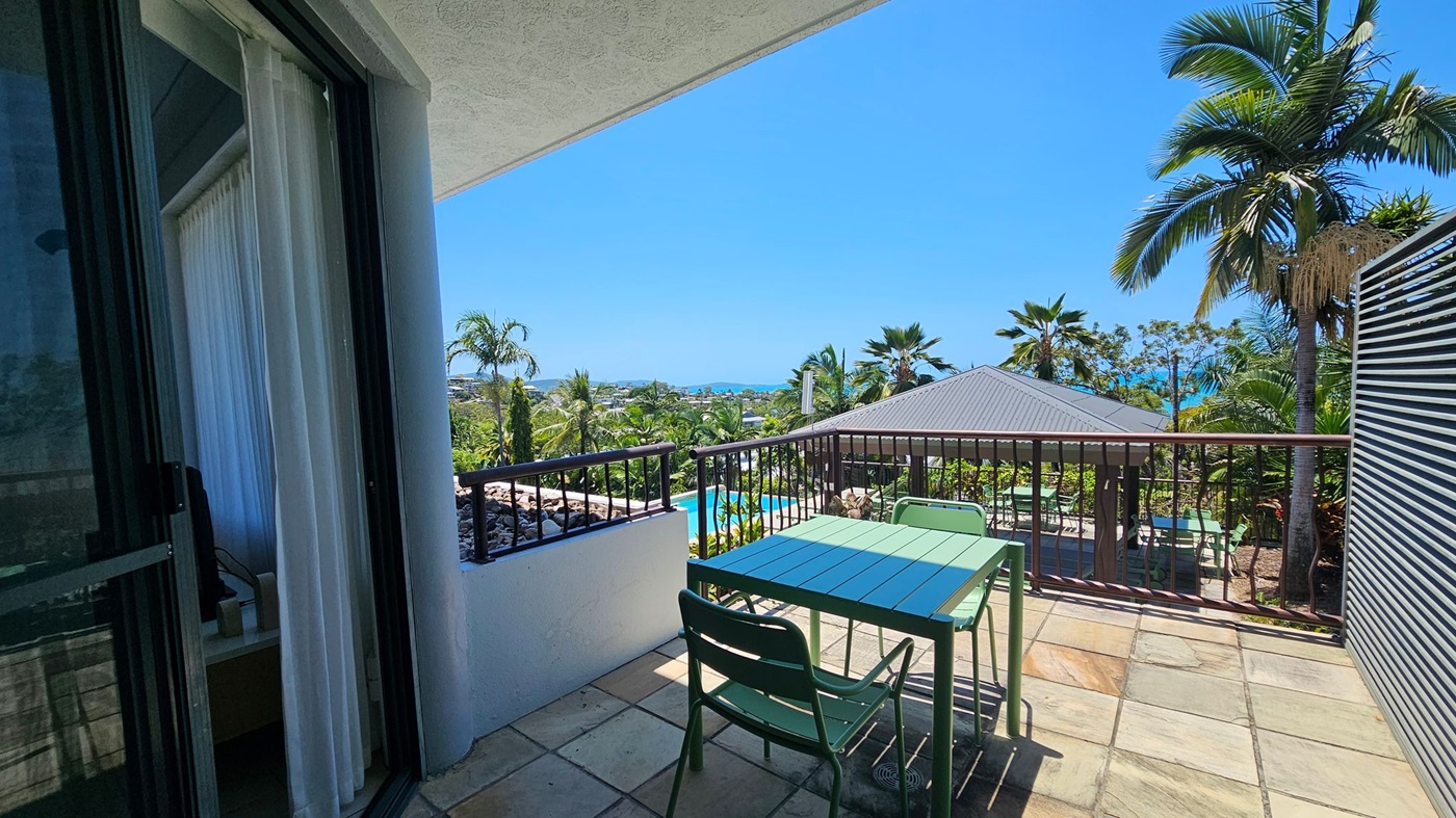 A balcony with a tiled floor features a green table and chairs. On the left, a sliding glass door with white curtains leads inside. The railing overlooks a lush garden with palm trees and a view of the ocean in the distance. A metal-roofed structure is visible on the right, surrounded by more greenery.