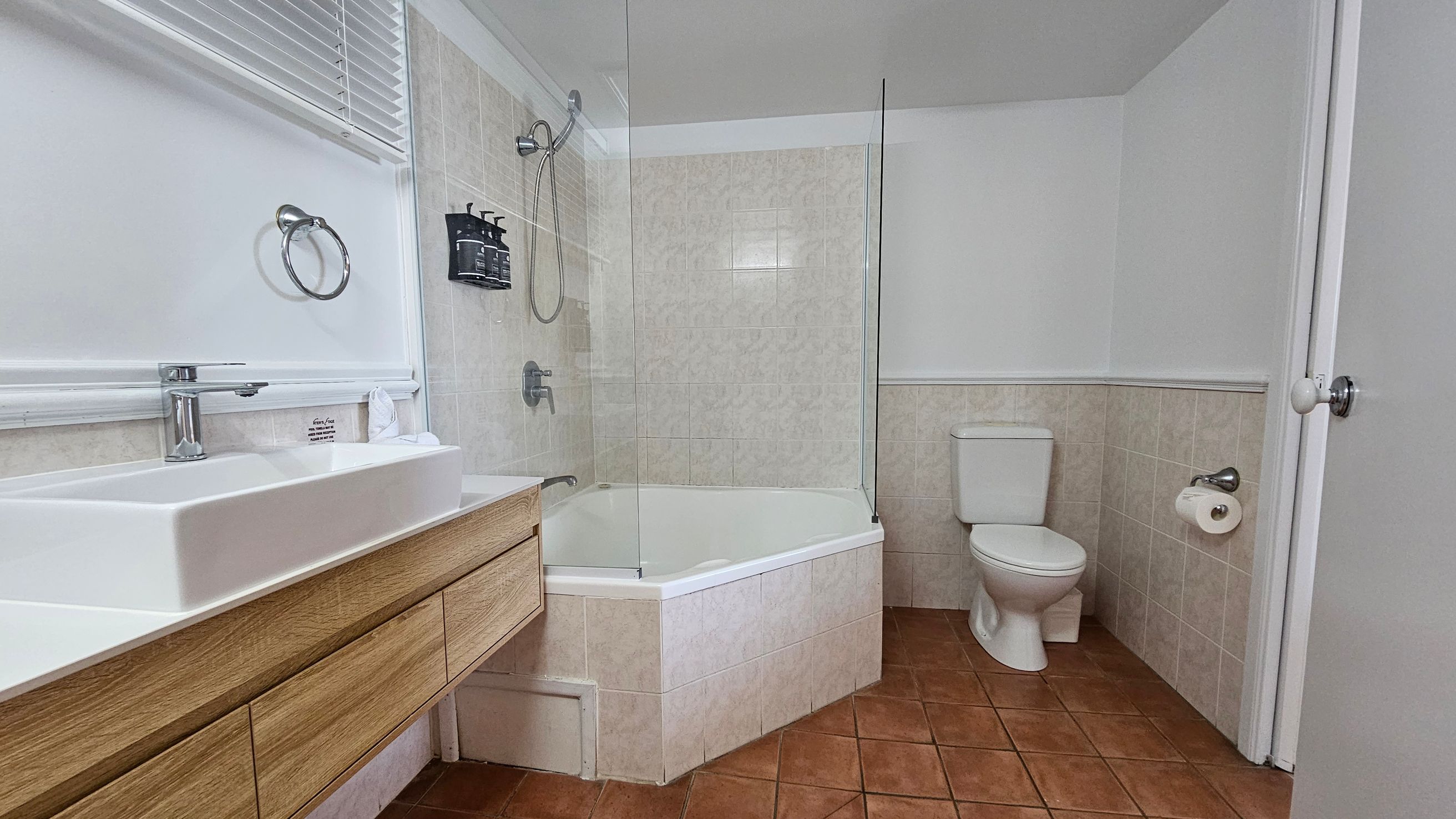 A bathroom with terracotta floor tiles and white walls. On the left, a wooden vanity with a white rectangular sink and a chrome tap is positioned beneath a window with white blinds. A towel ring is mounted on the wall. In the centre, a bathtub with a glass shower screen and wall-mounted showerhead is visible. On the right, a white toilet is next to a toilet roll holder. The ceiling is plain white, and a door with a round knob is partially open.
