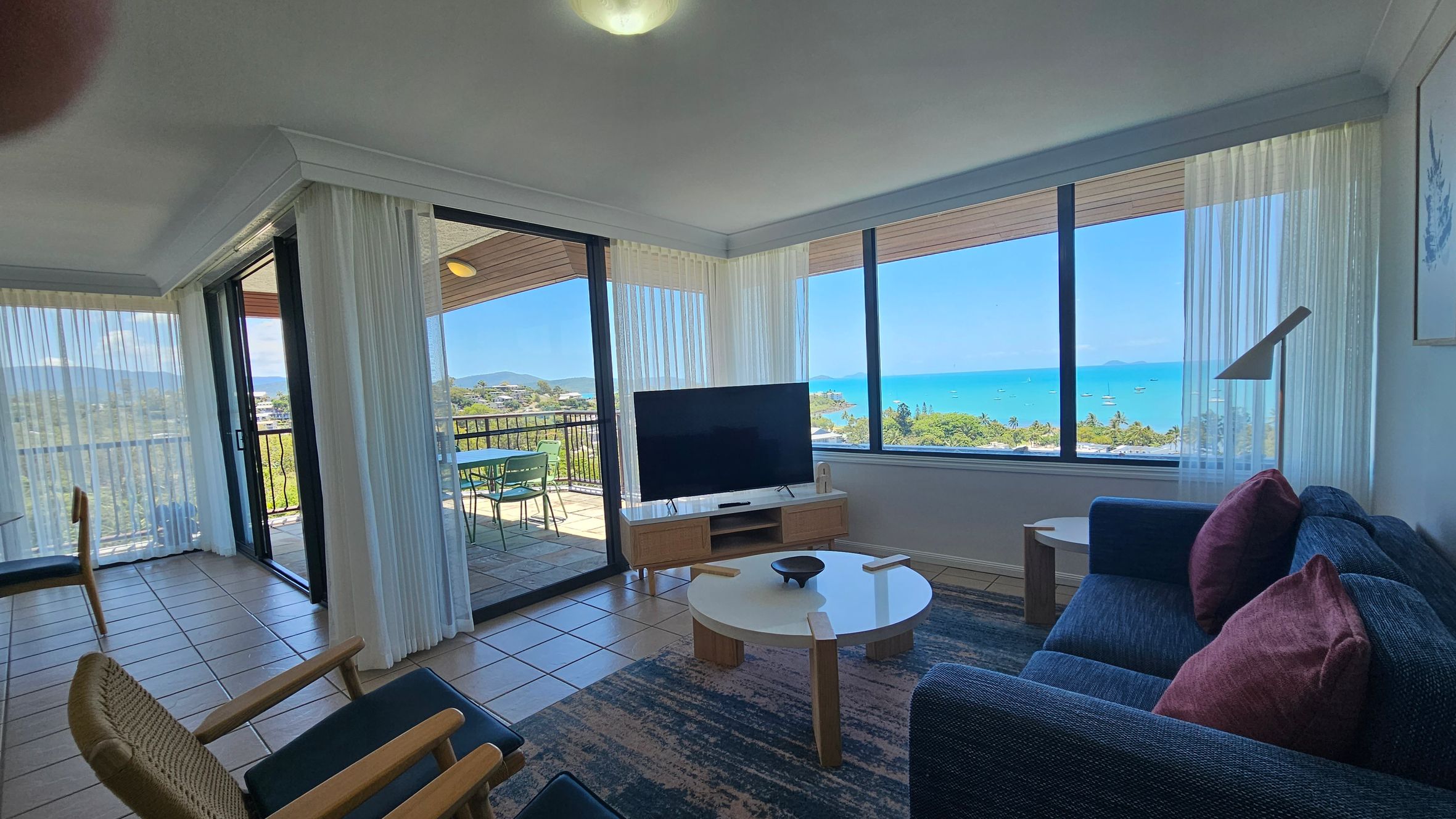 Open-plan living area with tiled flooring. On the left, a round dining table with wooden chairs is near sliding glass doors leading to a balcony with outdoor seating and a view of the sea. In the middle, a TV on a wooden stand faces a blue sofa with red cushions. On the right, a kitchen area with a white countertop and sink. The walls are adorned with framed artwork, and an air conditioning unit is mounted above. The ceiling has recessed lighting.