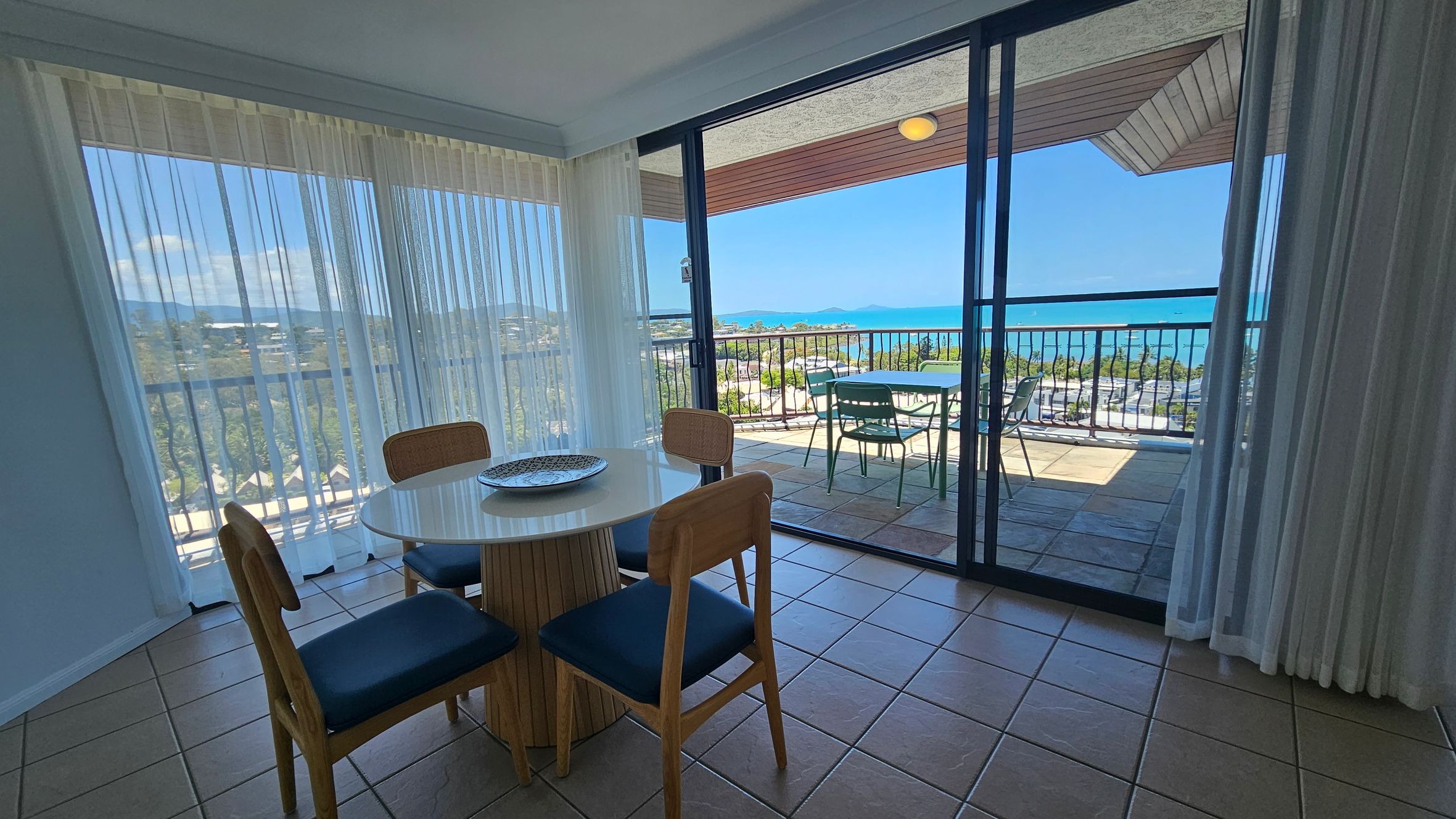 A room with a tiled floor features a round table with four wooden chairs, each with blue cushions, on the left. Sheer curtains cover large windows, offering a view of greenery and distant hills. On the right, sliding glass doors open to a balcony with a table and chairs, overlooking a scenic view of the ocean and coastline. The ceiling is plain with a light fixture above the balcony.