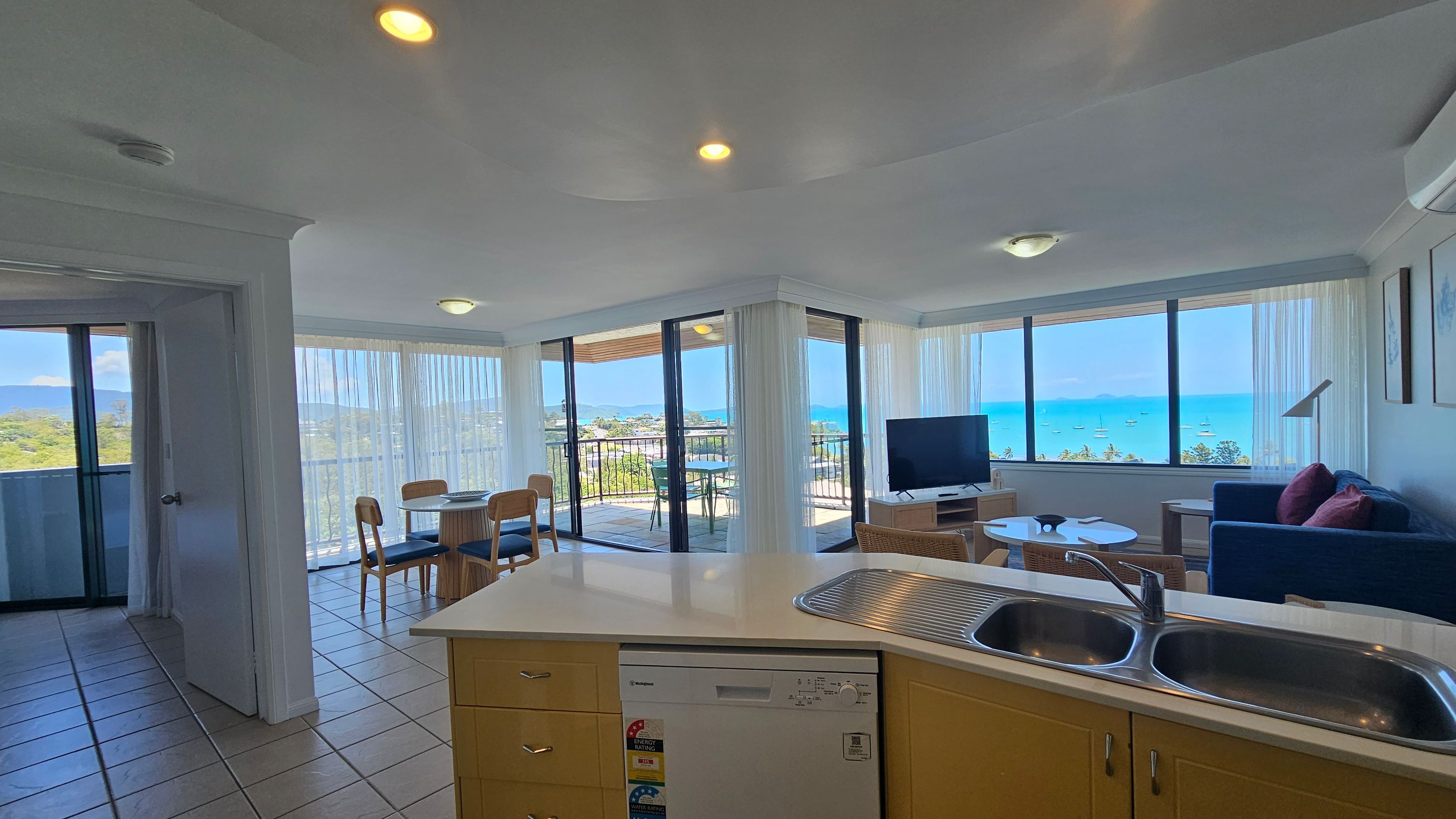 A bright, open-plan living space with a tiled floor. On the left, there's a dining area with a round table and chairs near large windows with sheer curtains. In the centre, a kitchen counter with a double sink and dishwasher. On the right, a seating area with a blue sofa, a TV on a stand, and a coffee table. Large windows and sliding doors lead to a balcony with views of the ocean and boats. The ceiling has recessed lighting.