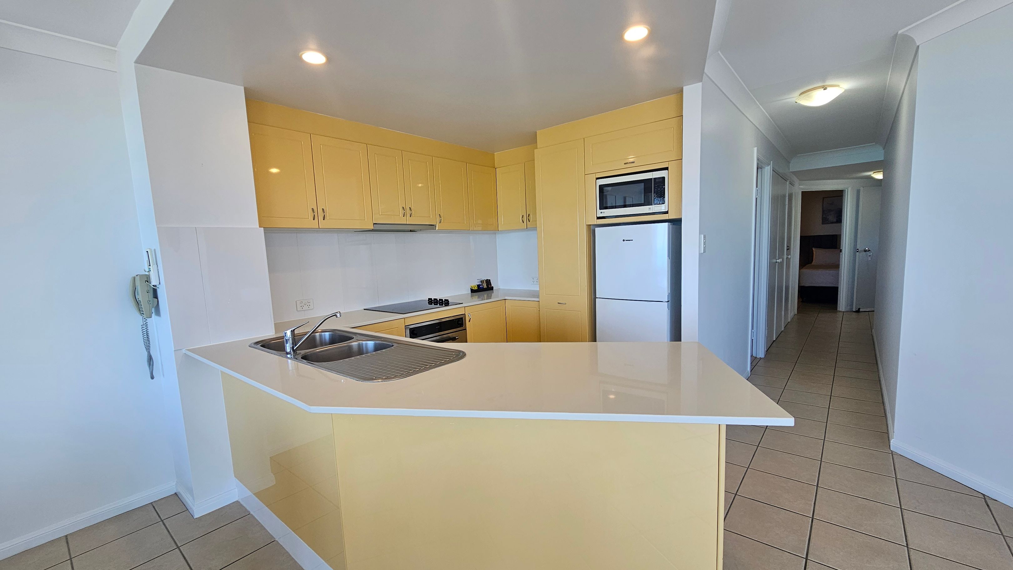 A kitchen with light yellow cabinets and a white countertop. On the left, there's a double sink with a draining board. The back wall has an oven, stovetop, and a microwave above a fridge. The floor is tiled in a light beige colour. To the right, a hallway leads to a room with a bed visible. The ceiling is white with recessed lighting.