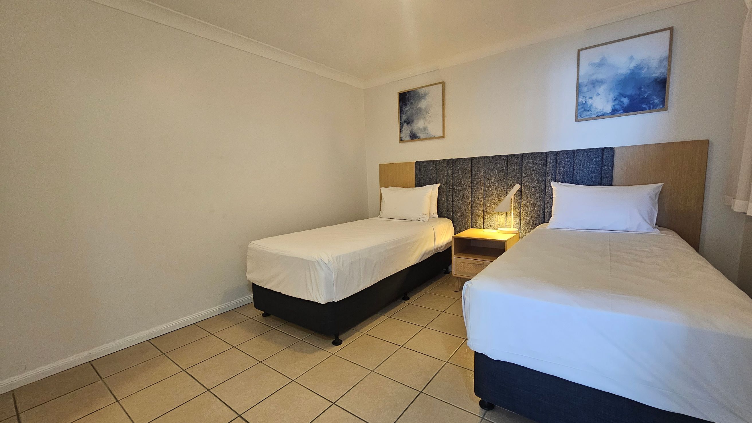 Two single beds with white bedding are positioned against a wall with a padded headboard. A small wooden nightstand with a lamp is between the beds. Above each bed hangs a framed abstract painting with blue tones. The floor is tiled in a light colour, and the walls are painted white. The ceiling is plain and white.