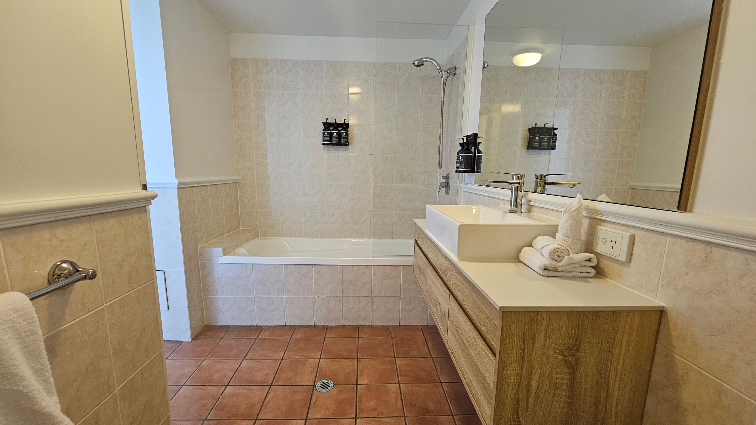 A bathroom with terracotta floor tiles and light-coloured wall tiles. On the left, a towel hangs on a rail. In the centre, a bathtub with a showerhead above and mounted dispensers on the wall. On the right, a wooden vanity with a white rectangular sink, chrome tap, and neatly folded towels. Above the sink is a large mirror reflecting the room. The ceiling is white with a round light fixture.