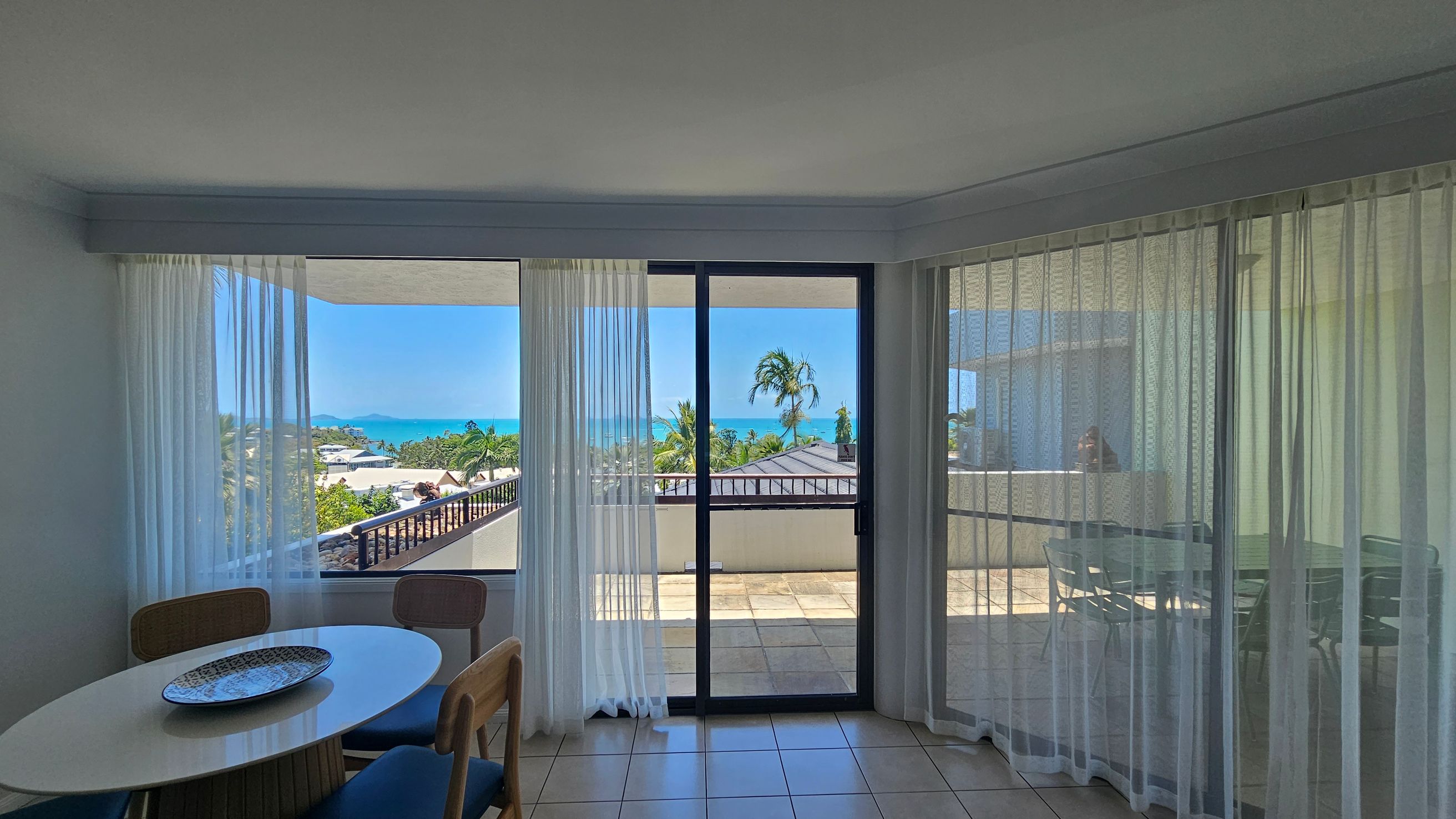 A room with large glass sliding doors and sheer curtains, offering a view of a balcony and a distant ocean. On the left, there's a round table with a decorative plate and several chairs. The balcony outside has a tiled floor and a railing, with palm trees and rooftops visible beyond. The floor inside is tiled, and the ceiling is plain white.