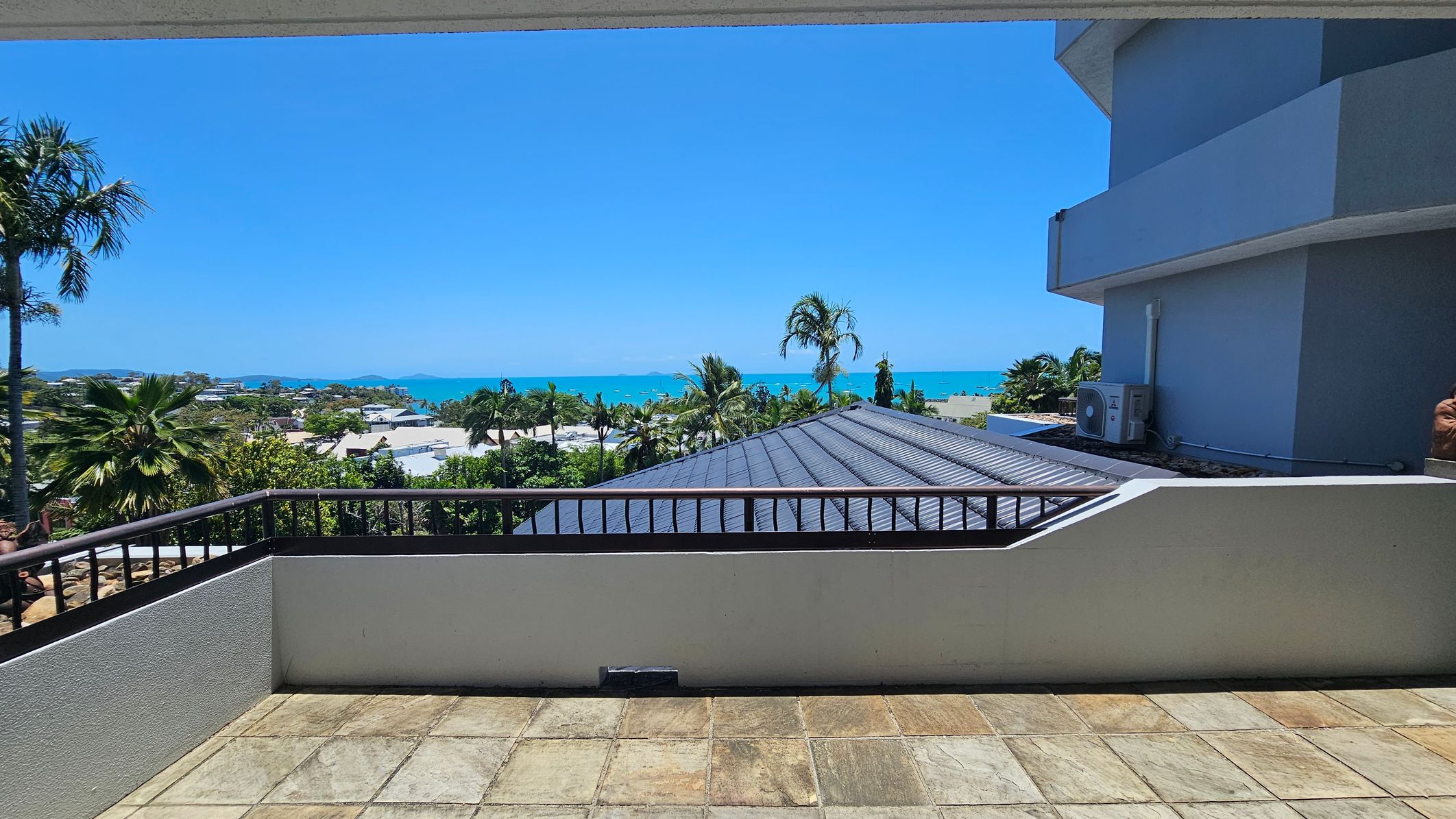 A terrace with a tiled floor overlooks a scenic view. In the foreground, a low wall with a railing runs along the edge. To the left, palm trees and lush greenery are visible. In the centre, rooftops and more palm trees lead to a distant view of the sea. On the right, part of a modern building with an air conditioning unit is visible. The background features a clear blue sky and the ocean.