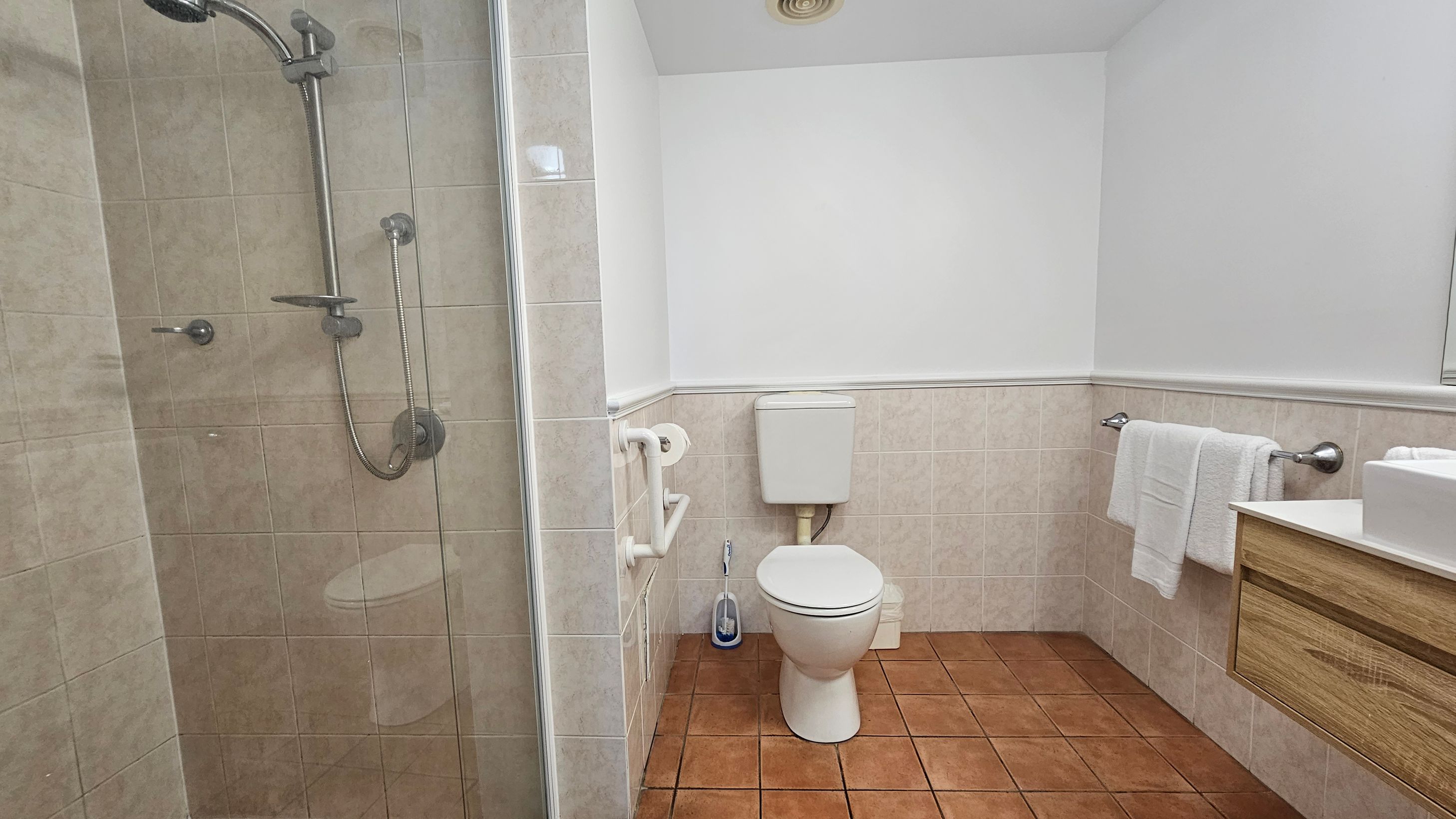 A bathroom with a tiled floor and walls. On the left, there is a glass-enclosed shower with a silver showerhead and controls. In the centre, a white toilet with a cistern is mounted on the wall, accompanied by a toilet brush and holder. A white grab bar and toilet roll holder are next to the toilet. On the right, a wooden vanity with a white basin and a towel rail holding white towels. The ceiling is plain white with a vent.