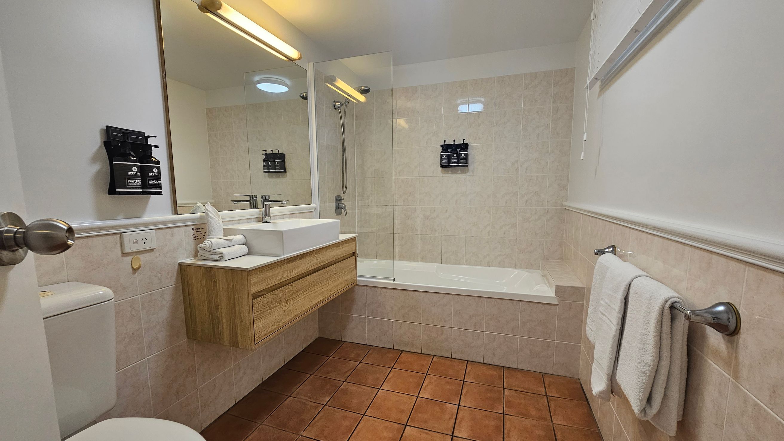 A bathroom with terracotta floor tiles and beige wall tiles. On the left, there's a toilet and a wall-mounted soap dispenser. A wooden vanity with a white basin and a mirror above it is in the centre. On the right, a towel rail holds two white towels. The bathtub with a glass screen is along the back wall, featuring a showerhead and another soap dispenser. The ceiling has a light fixture.