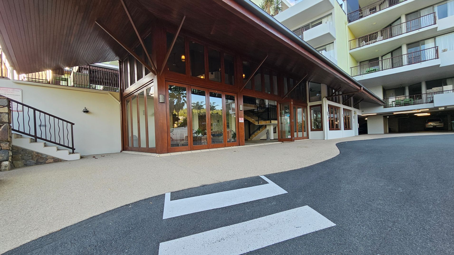 A building entrance with large wooden-framed glass doors is in the centre. To the left, a staircase with a black metal railing leads up, adjacent to a stone wall. The ceiling is wooden and sloped. On the right, a multi-storey building with balconies and a covered parking area is visible. The foreground features a paved area with a zebra crossing on the asphalt road.