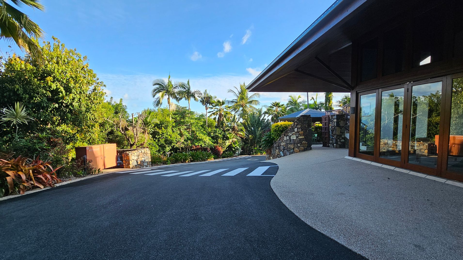 A paved road with a zebra crossing leads to a building on the right, featuring large glass doors and a stone wall. Lush greenery with palm trees and various plants fills the left side and background. A stone structure and a brown utility box are visible on the left. The foreground has a smooth, light-coloured pavement adjacent to the road.