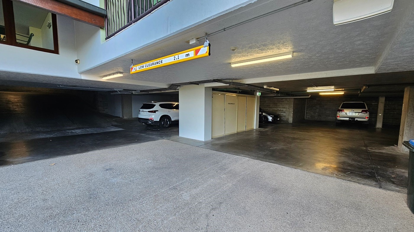 Underground car park with a low clearance sign of 2.1 metres hanging from the ceiling. On the left, there is a dark, empty parking space. In the centre, a white SUV is parked next to a storage unit. On the right, another vehicle is parked near a concrete wall. The floor is a mix of concrete and asphalt, and the ceiling is low with fluorescent lights. A staircase is visible in the upper left corner.