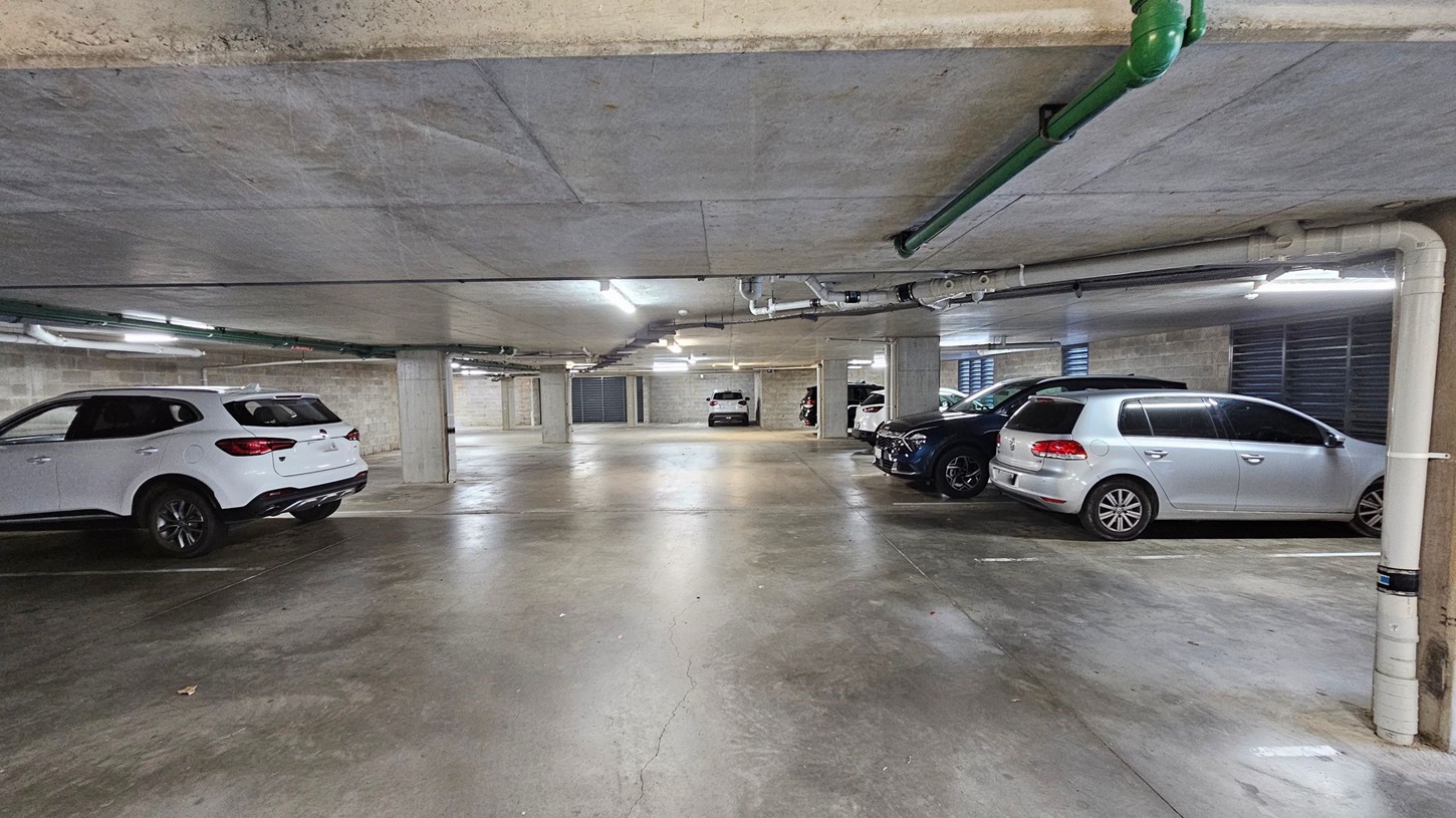 A concrete underground car park with several parked cars. On the left, a white SUV is parked near a wall. On the right, a silver hatchback and a dark-coloured SUV are parked side by side. The floor is concrete with some greenish patches. The ceiling is low with exposed pipes and fluorescent lights. In the background, more cars are visible, and there are concrete pillars throughout the space.