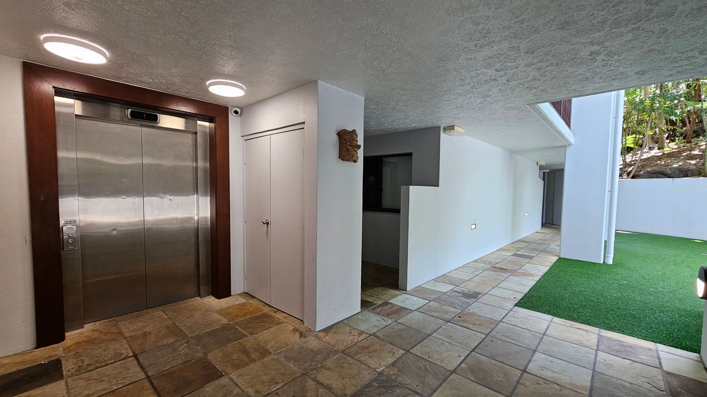 On the left, there is a stainless steel lift with a wooden frame, illuminated by two circular ceiling lights. Next to the lift is a small white door. The floor is tiled with a mix of earthy tones. On the right, a corridor leads to a door, with a section of artificial grass adjacent to it. The walls are white, and the ceiling has a textured finish. In the background, there is a glimpse of greenery through an open area.