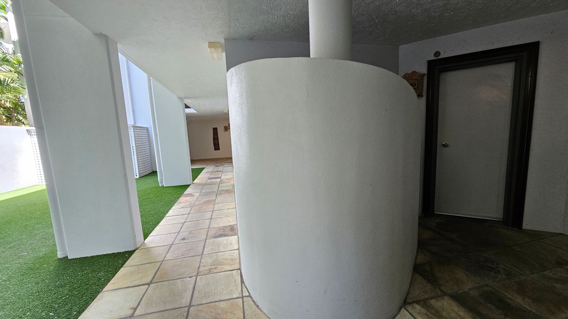 A corridor with a tiled floor and a white ceiling. On the right, there's a curved white wall and a door with a dark frame. On the left, there are white pillars and a patch of artificial grass. In the background, more tiles and a beige wall with a decorative piece.