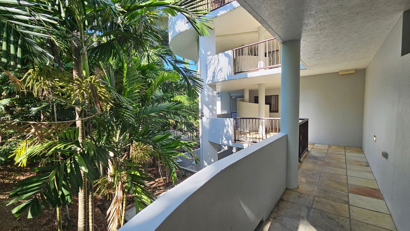On the left, there are lush green palm trees with dense foliage. On the right, a building with white walls and multiple balconies featuring metal railings. The floor is tiled with a mix of green and brown tiles, and the ceiling is smooth and light-coloured. The scene is well-lit, suggesting daylight.