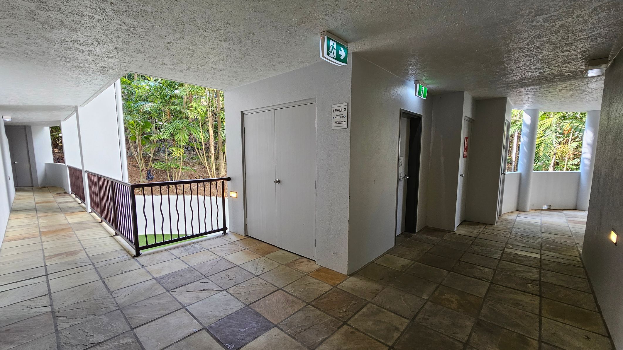A corridor with a tiled floor and a textured ceiling. On the left, there's a railing with a view of lush green trees outside. In the centre, a white door with a sign indicating "Level 2" and an emergency exit sign above. On the right, two doors are visible, with more greenery seen through windows. The corridor is well-lit with wall lights and natural light from the windows.