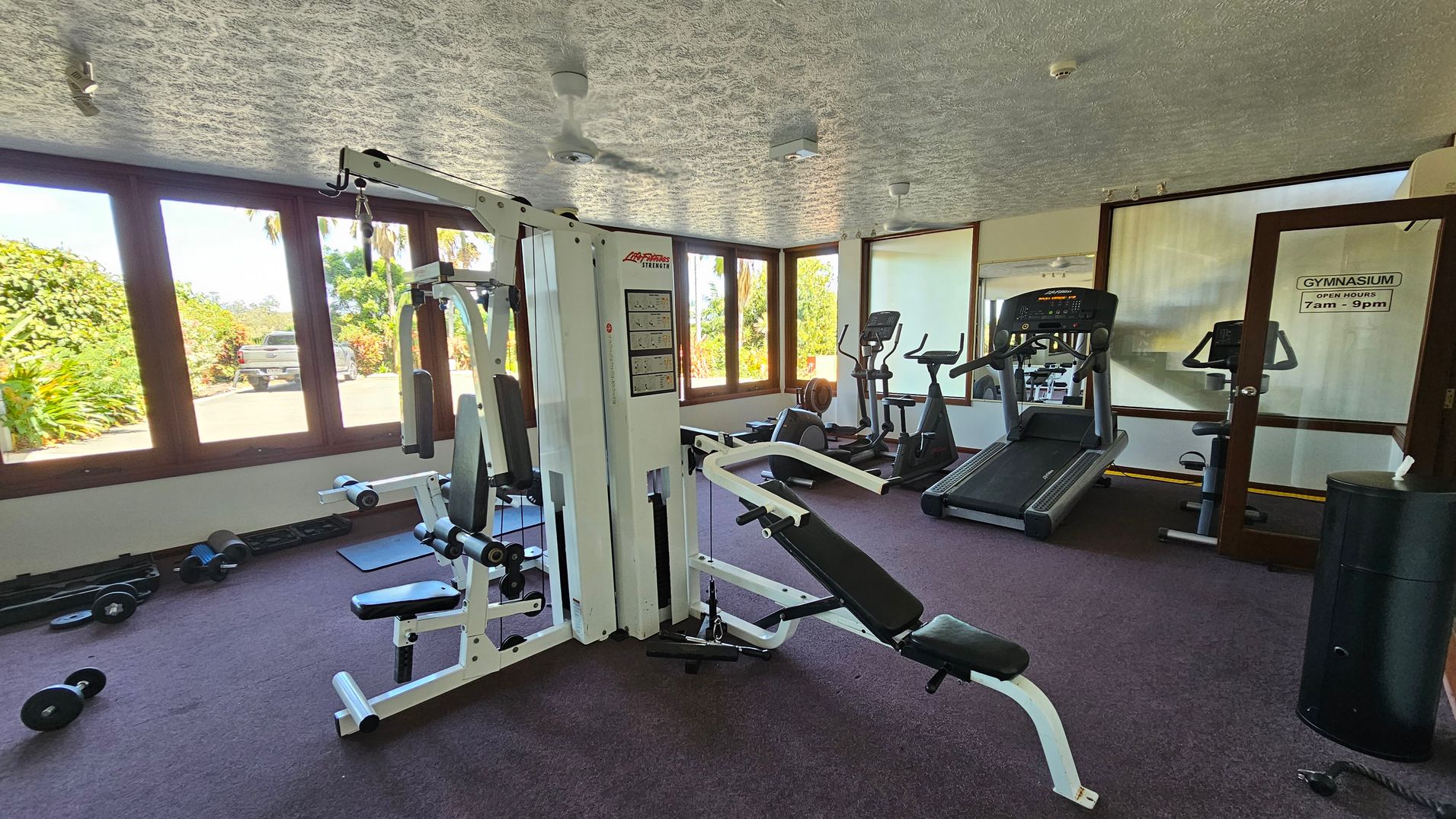 A gym with various exercise equipment. On the left, there are dumbbells on the floor and a multi-gym machine. In the middle, a bench press is positioned. On the right, there are treadmills and an exercise bike near a glass door with a sign indicating gym hours. Large windows in the background reveal greenery and a parked vehicle outside. The ceiling is textured, and the floor is carpeted.