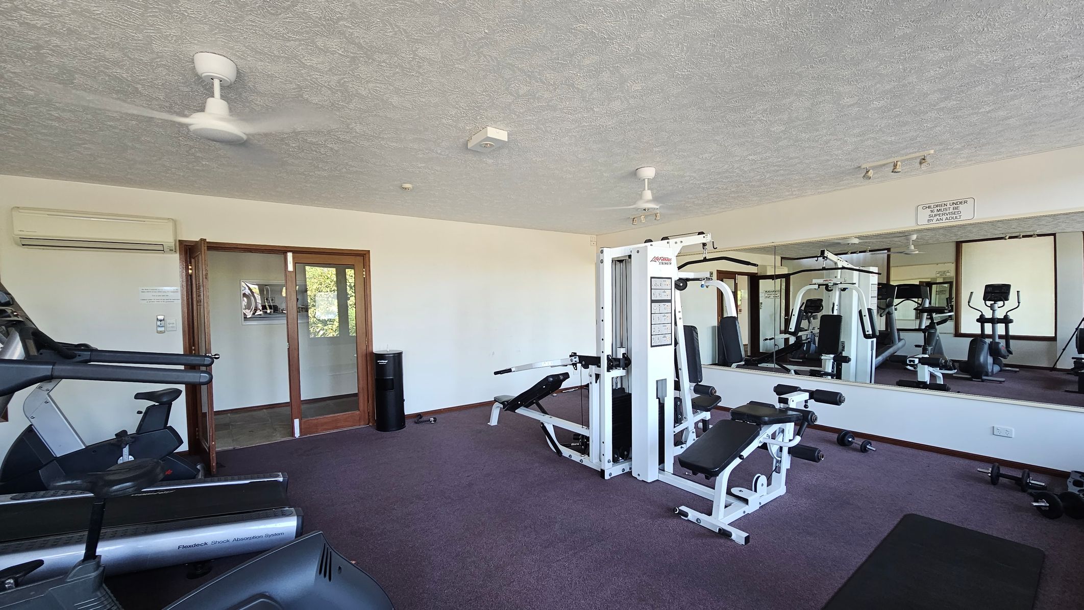 A gym room with various exercise equipment. On the left, there are treadmills and stationary bikes. In the centre, a white multi-gym machine is positioned. The right side features a large mirror reflecting the equipment and a sign about supervision. The floor is carpeted in dark grey, and the ceiling is textured with two ceiling fans. A wooden-framed glass door is on the far wall, and an air conditioning unit is mounted above it.
