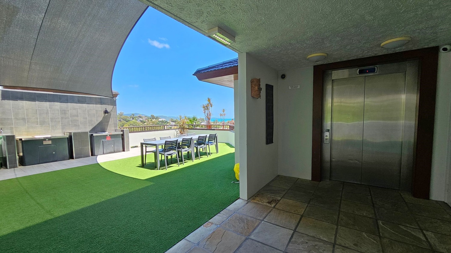 On the right, there is a metallic lift door set in a tiled wall. The floor in front of the lift is made of square tiles. On the left, a shaded outdoor area features a green artificial grass floor, a long table with several chairs, and a barbecue setup against a tiled wall. In the background, there is a view of distant hills and the sea, with a few small trees and a railing along the edge of the terrace.