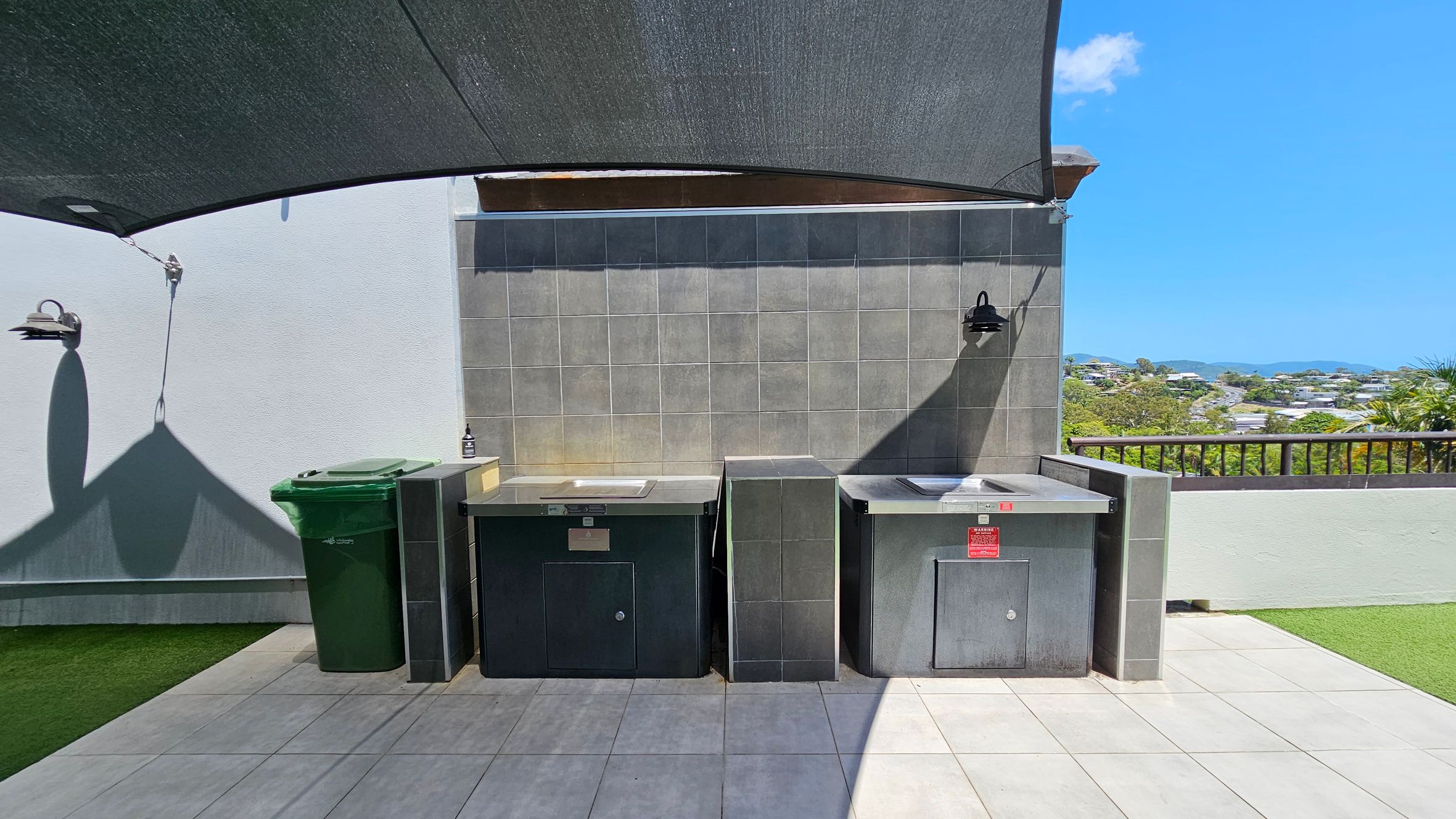 Outdoor area with a tiled floor and a grey tiled wall in the background. Two stainless steel barbecue units are positioned side by side against the wall. A green rubbish bin is on the left. A black shade cloth is overhead, casting shadows on the wall. Two black wall-mounted lights are above the barbecues. To the right, a railing overlooks a view of trees and houses in the distance. The area is bordered by artificial grass.