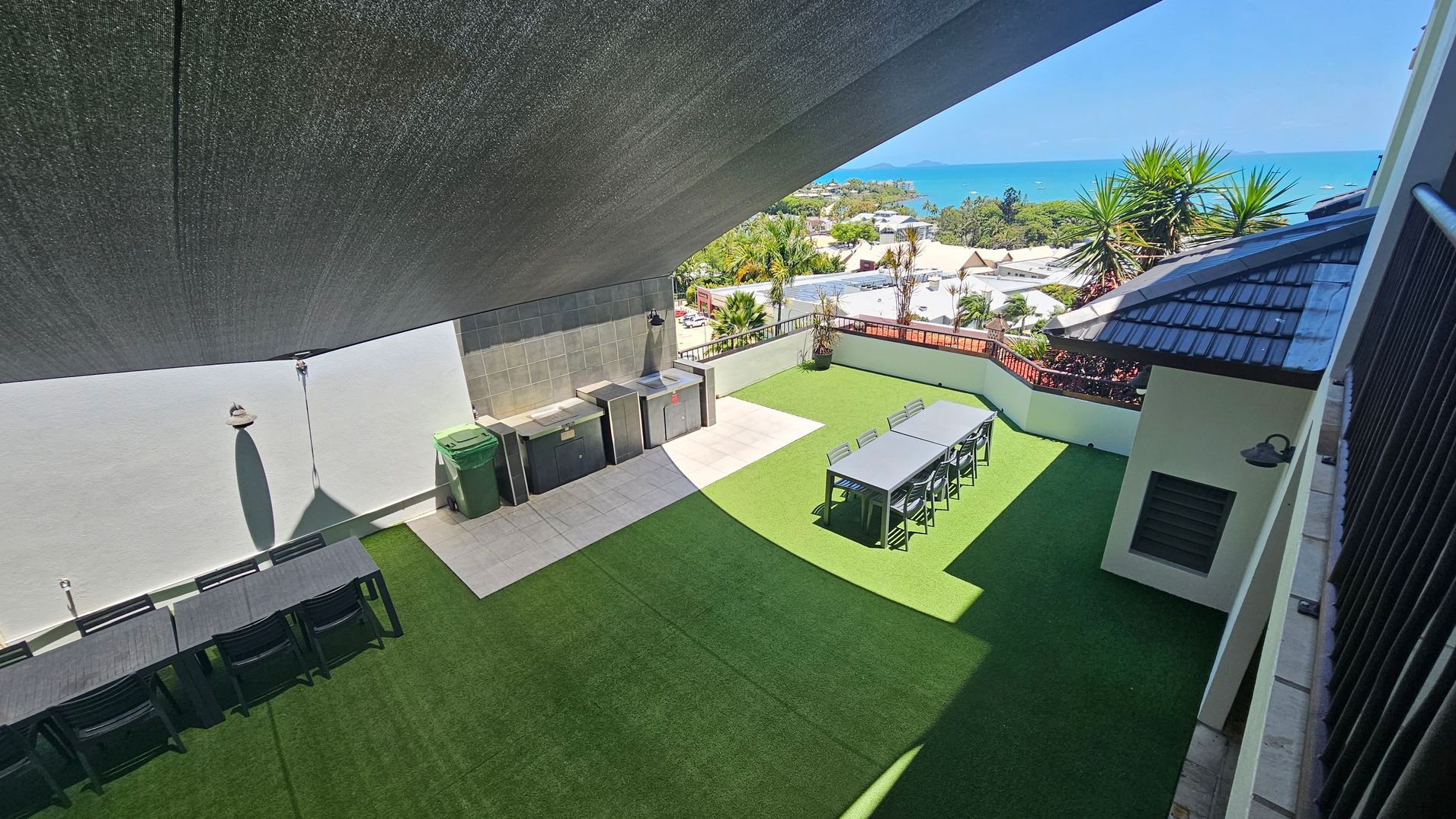 A rooftop terrace with artificial grass flooring. On the left, there is a long table with chairs and a wall-mounted lamp casting a shadow. In the middle, a tiled area features a sink and storage units, with a green bin nearby. On the right, another table with chairs is positioned near the edge. The background shows a view of the sea, palm trees, and rooftops. A large shade cloth covers the area above.