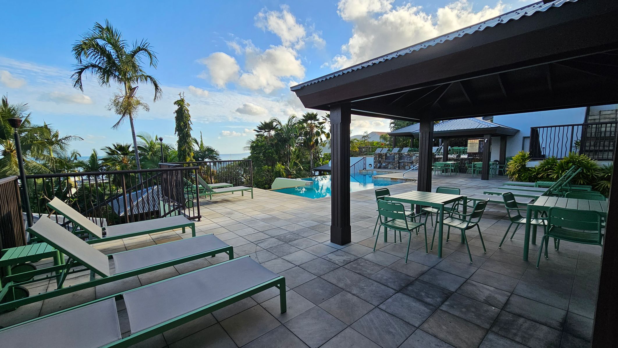 A poolside area with tiled flooring. On the left, there are several green sun loungers and a railing with tropical plants and palm trees beyond. In the centre, an infinity pool is visible, surrounded by more greenery. On the right, there is a covered seating area with green tables and chairs. A building with a metal roof is in the background, and more seating is visible near the pool.