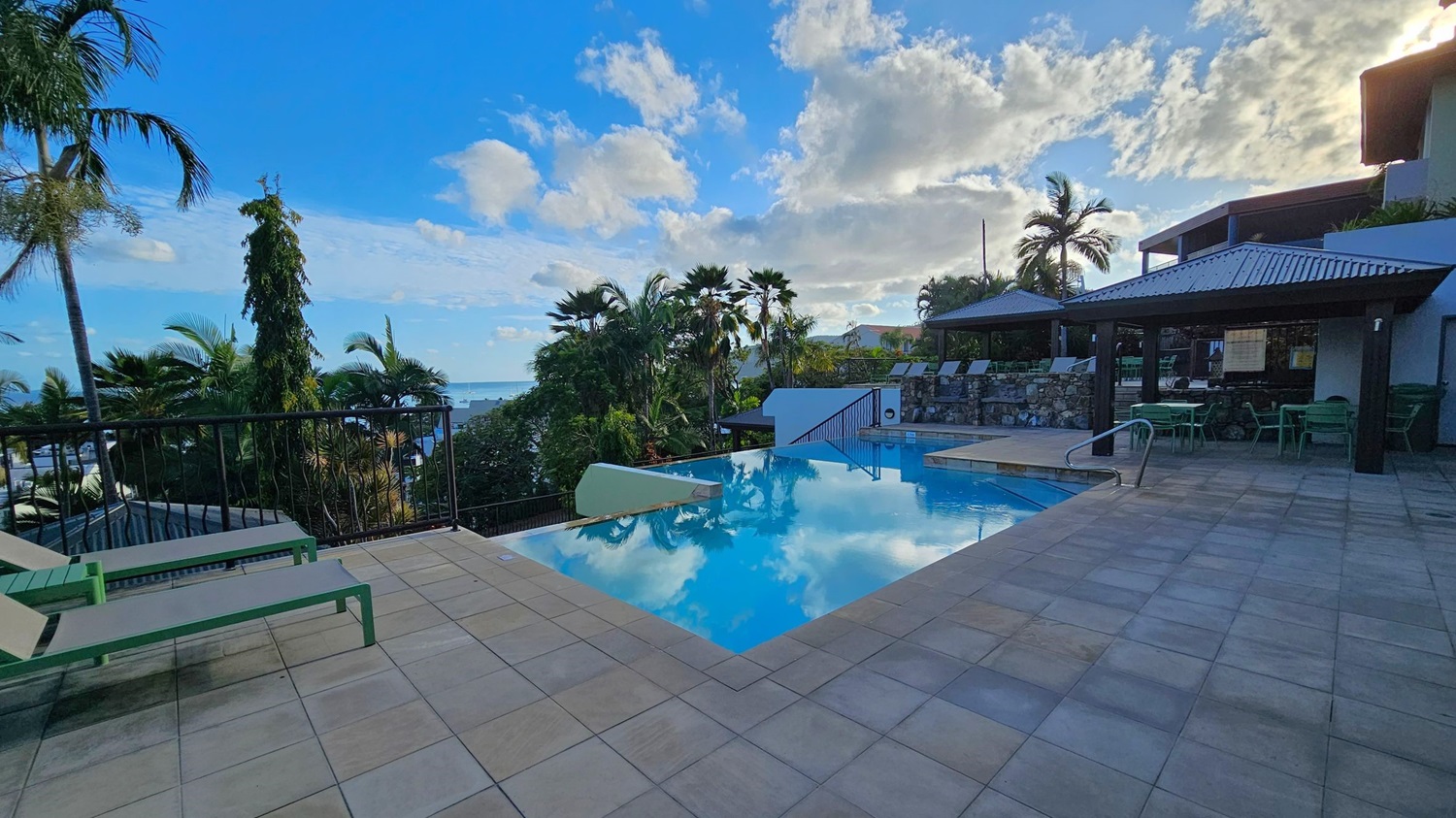 A serene pool area with a tiled deck. On the left, there are two green sun loungers. The centre features an infinity pool reflecting the sky and clouds. To the right, a covered seating area with green chairs and tables is visible. In the background, lush palm trees and foliage surround the area, with a glimpse of the sea and boats in the distance.