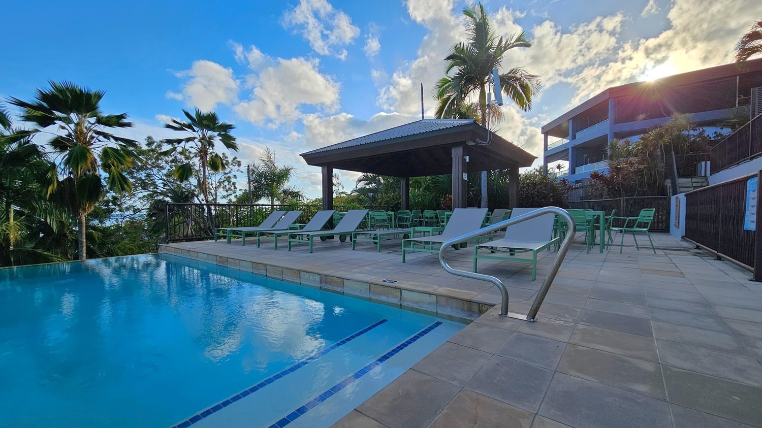 An outdoor pool with clear blue water is on the left, bordered by a tiled deck. On the right, several sun loungers and green chairs are arranged under a wooden gazebo. Palm trees are scattered around, and a modern building is visible in the background. The sun is partially hidden behind the building, casting light over the scene.