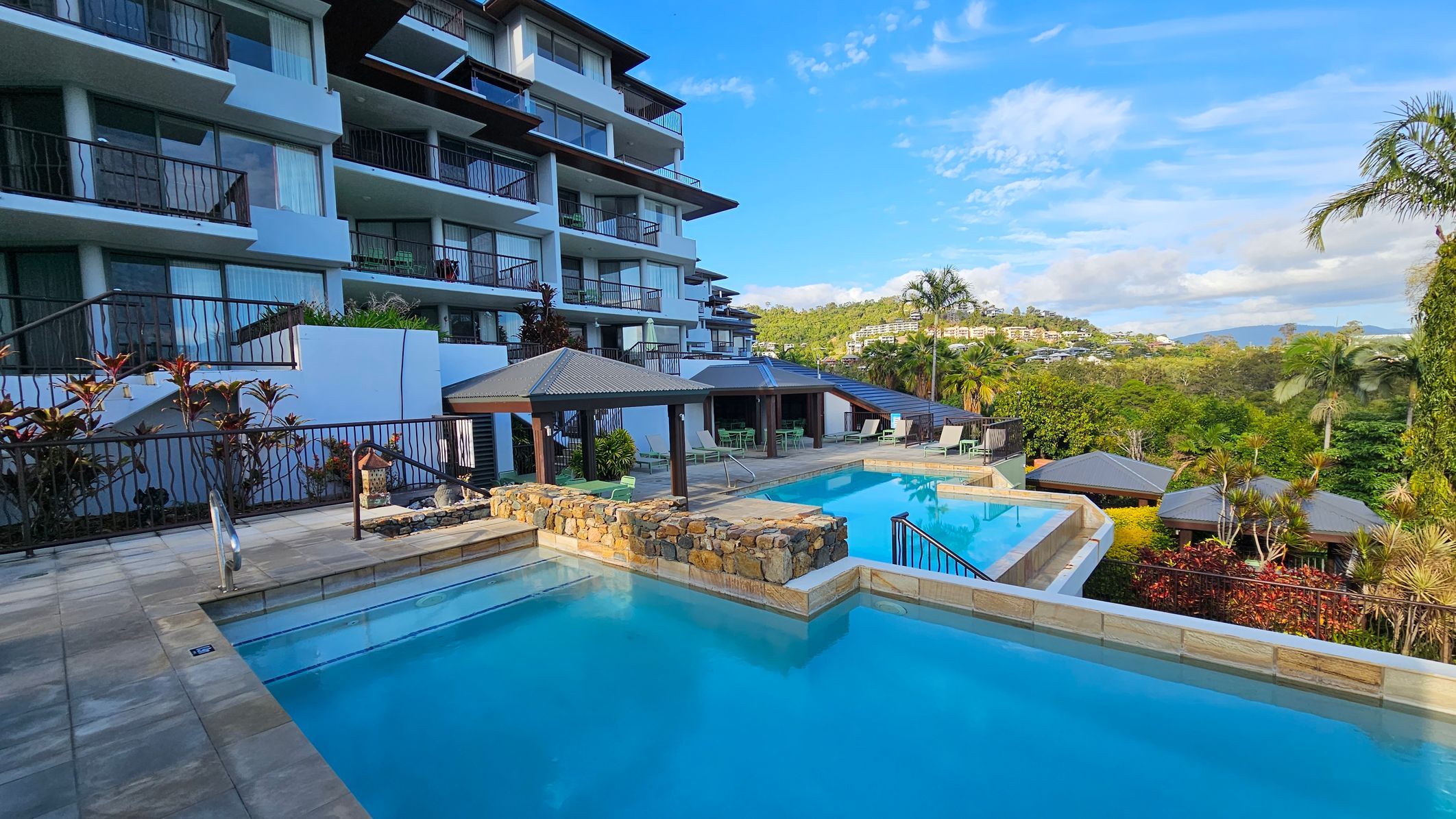 A multi-storey building with balconies is on the left, featuring glass doors and railings. In the centre, there are two swimming pools separated by a stone wall, with a tiled deck surrounding them. Several shaded seating areas with tables and chairs are near the pools. On the right, lush greenery and palm trees are visible, with hills and more buildings in the distance.