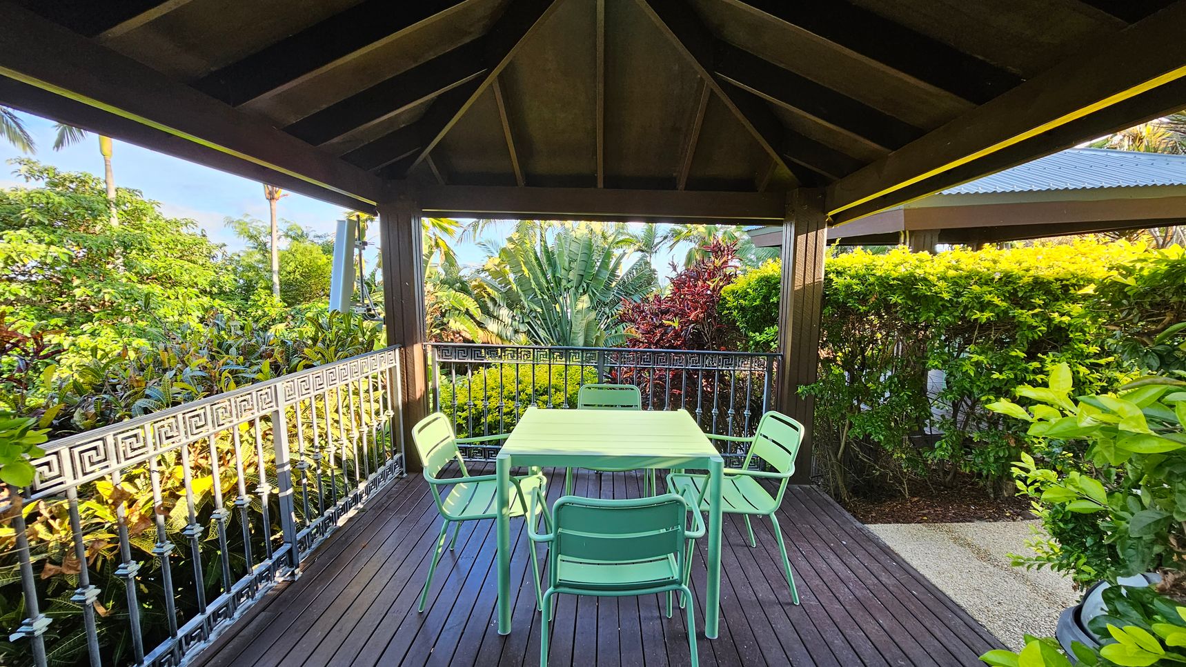 A covered patio area with a small pool in the foreground. The floor is tiled in a light stone pattern. To the left, a column with "push to start" and "stop" buttons. In the middle, a green table with matching chairs and two white sun loungers. The background features lush tropical plants and trees, with a metal railing separating the patio from the garden. The ceiling is dark wood with exposed beams.