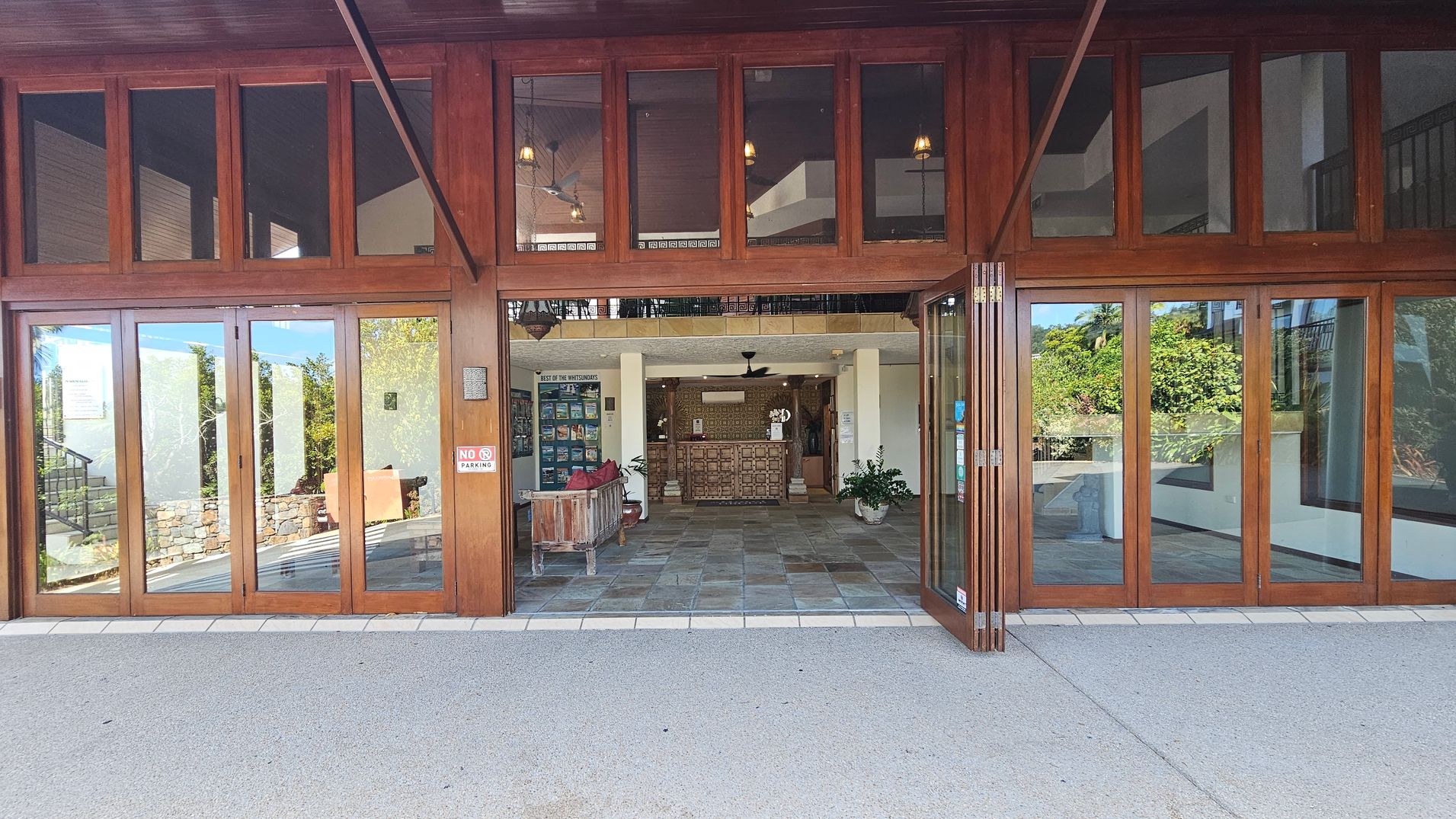A building entrance with large wooden-framed glass doors. Inside, a reception area with a wooden desk is visible. To the left, a wooden bench with red cushions and a display of brochures. To the right, a potted plant. The floor is tiled with a mix of earthy tones. The ceiling has a fan and hanging lights. Outside, a "No Parking" sign is on the door. The background shows greenery through the glass.