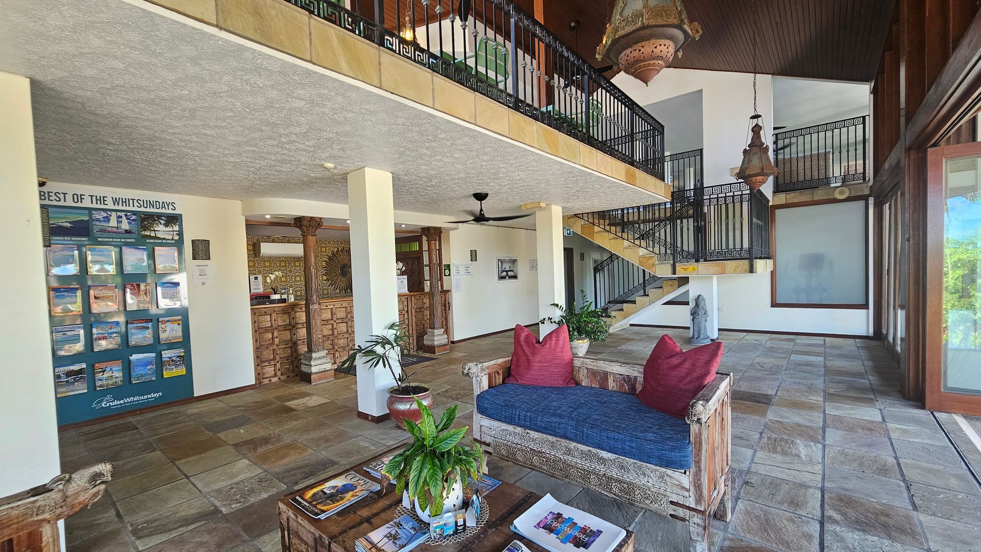 A spacious interior with a stone-tiled floor and a textured ceiling. On the left, a display board titled "Best of the Whitsundays" features brochures. In the centre, a wooden reception desk with ornate columns is visible. A rustic wooden bench with red cushions and a blue seat is in the foreground, accompanied by a wooden table with magazines and a plant. A staircase with black railings leads to an upper level. Large windows on the right allow natural light to enter.