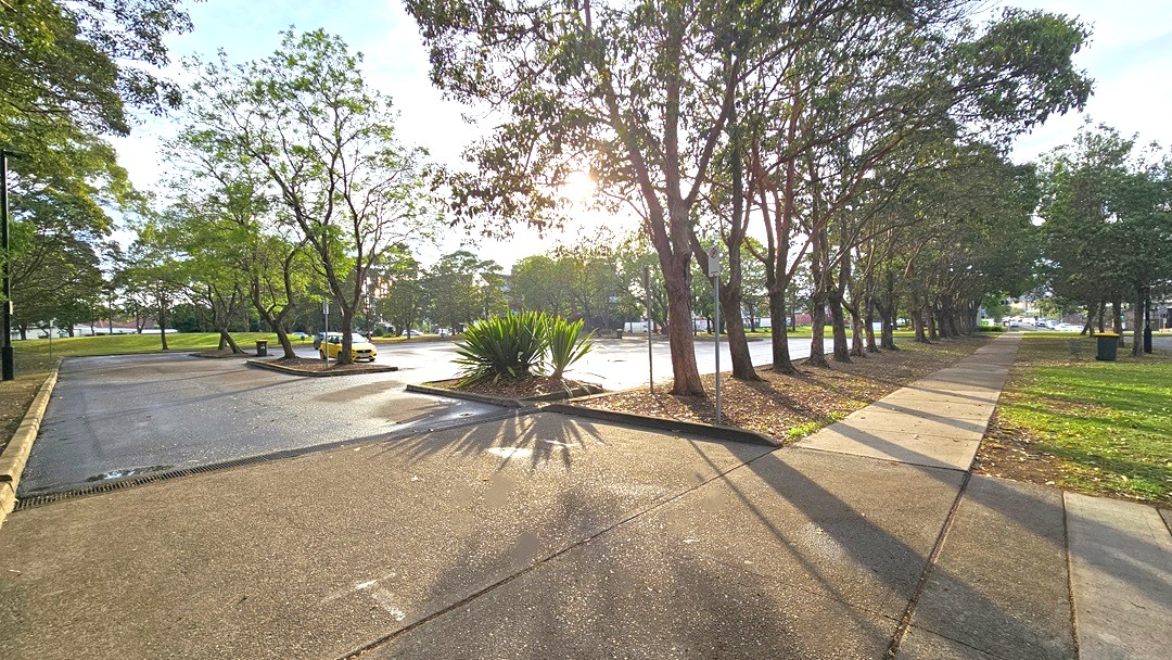 Extensive car park from the right side of the facility. Around the car park are trees lined on the edge of the garden. A No Parking sign with a red arrow at the bottom pointing towards the entry space to the extensive car park can be found on the right of the main entrance. Near it are two green plants from the palm family. Large grey trash bins with yellow covers are placed inside the car park and on the side of the concrete walkway.
